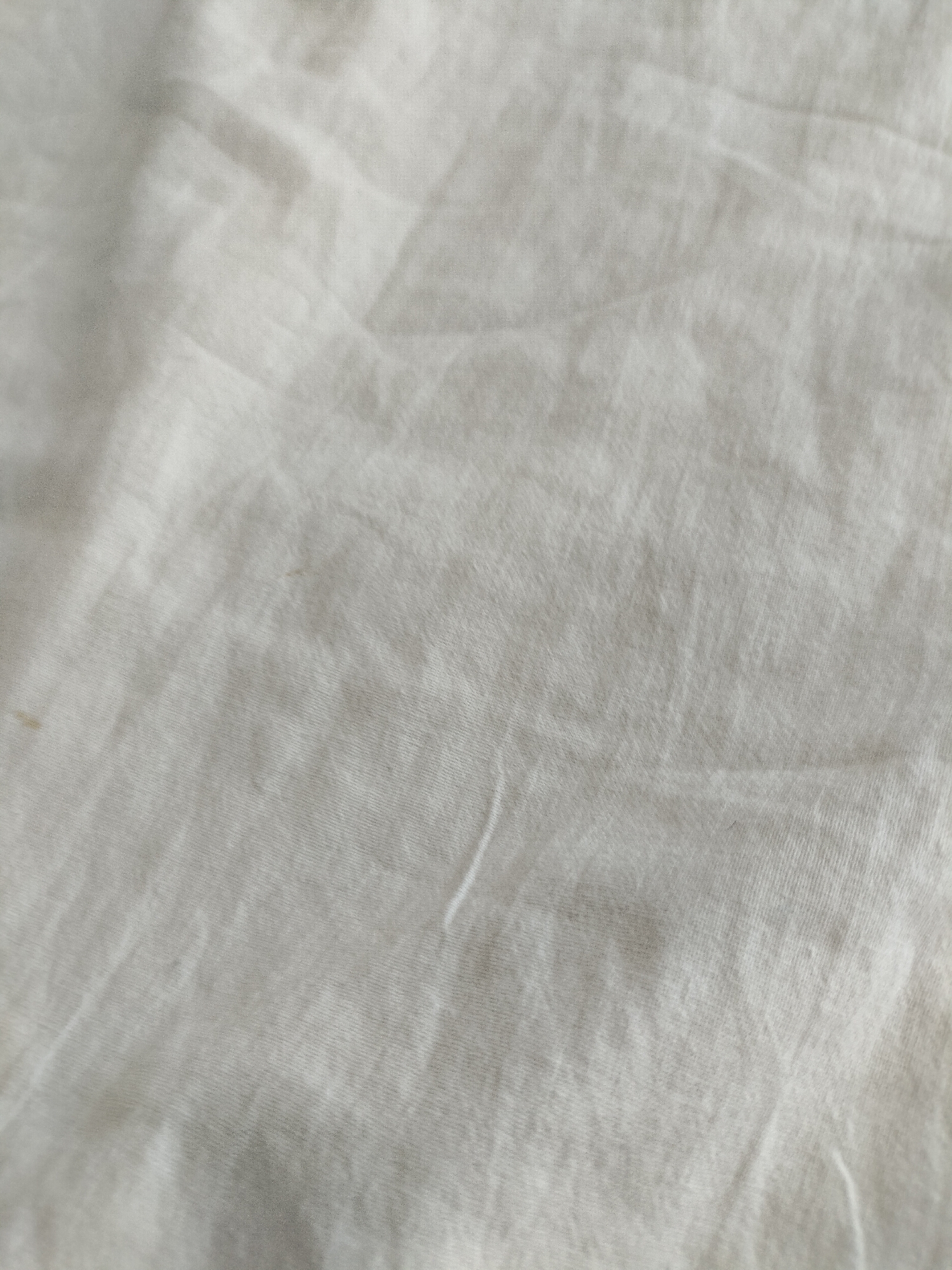 The image is a close-up, overhead view of a wrinkled, light-colored fabric. The fabric appears to be cotton or a similar material, with a visible weave pattern. There are subtle shadows and highlights created by the folds and creases in the material, suggesting it is not perfectly flat. Two small, faint brown stains are visible on the fabric in the lower left portion of the image. No people, objects, or environmental features beyond the fabric itself are discernible. The image provides no information regarding its location in Chernihiv, Ukraine, or any other geographical context.