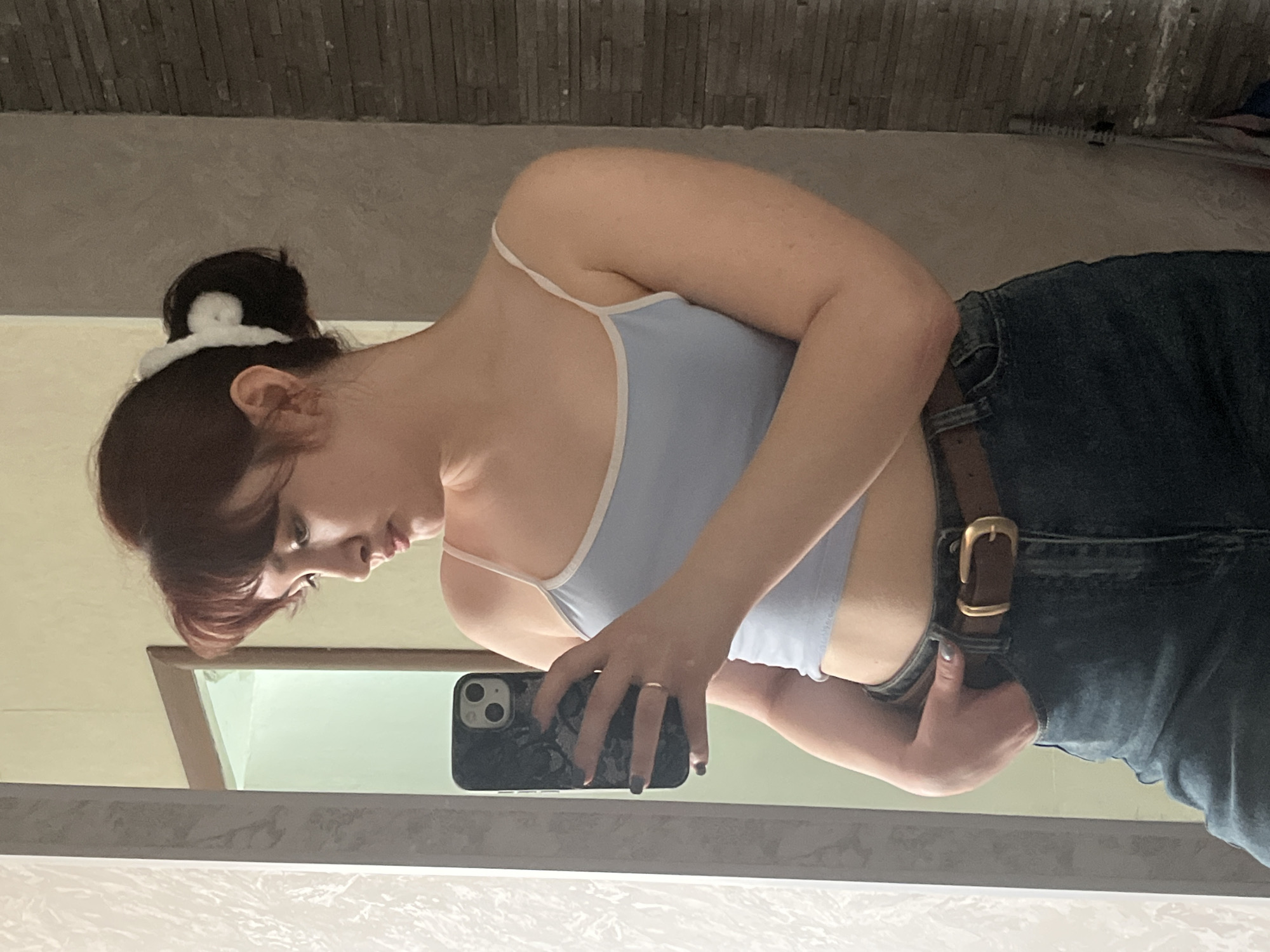 A mirror-reflection selfie captures a young woman in a light blue tank top and dark blue jeans secured with a brown leather belt. Her hair is pulled back into a bun with a white scrunchie. She holds a black smartphone with both hands, taking the photo in front of a bathroom mirror. The bathroom's décor includes gray brick-patterned walls and a white ceiling. The location is not discernible from the image and cannot be confirmed as Neftekamsk, Russia.