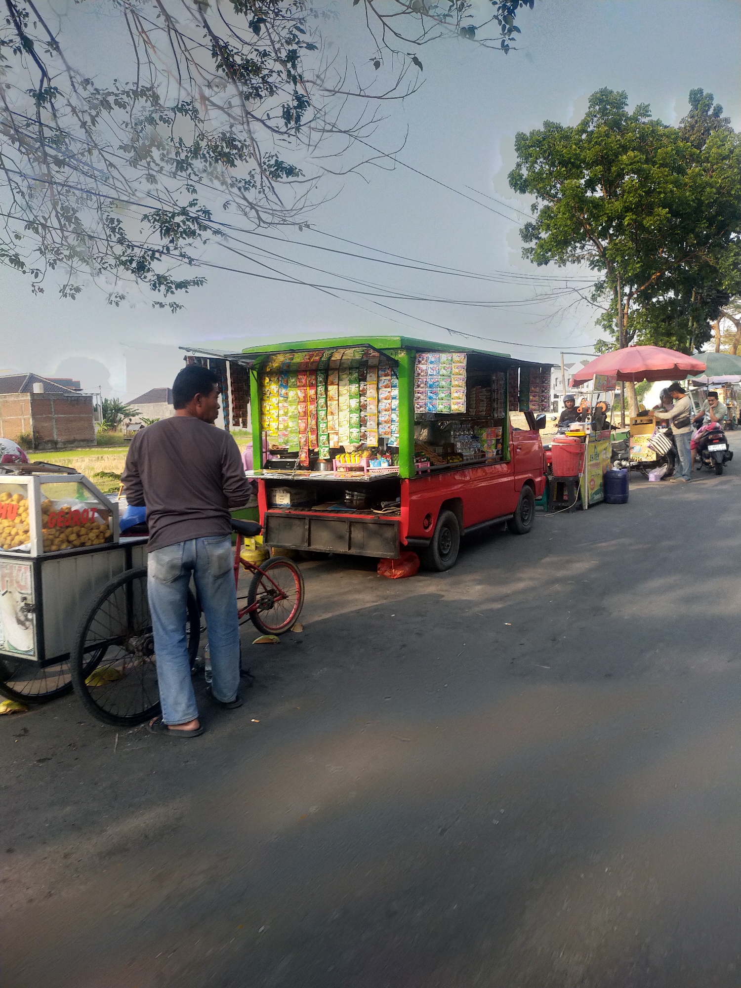 This is a daylight, eye-level, medium shot of a street scene in Slawi, Indonesia, capturing a row of roadside food stalls and vendors. The primary focus is a red pickup truck, converted into a mobile store, stocked with an assortment of packaged snacks displayed on its sides. To the left of the truck, a man in a dark long-sleeved shirt and blue jeans stands beside a cart filled with fried food items, with a red bicycle leaning against it. Further down the street, other vendors are visible, including one operating under a red umbrella. Several motorcycles are parked nearby, indicating potential customer activity. The background shows residential buildings and a lush green tree, with overhead utility wires crisscrossing the sky. The overall setting suggests a typical market or roadside commercial area in an Indonesian city.