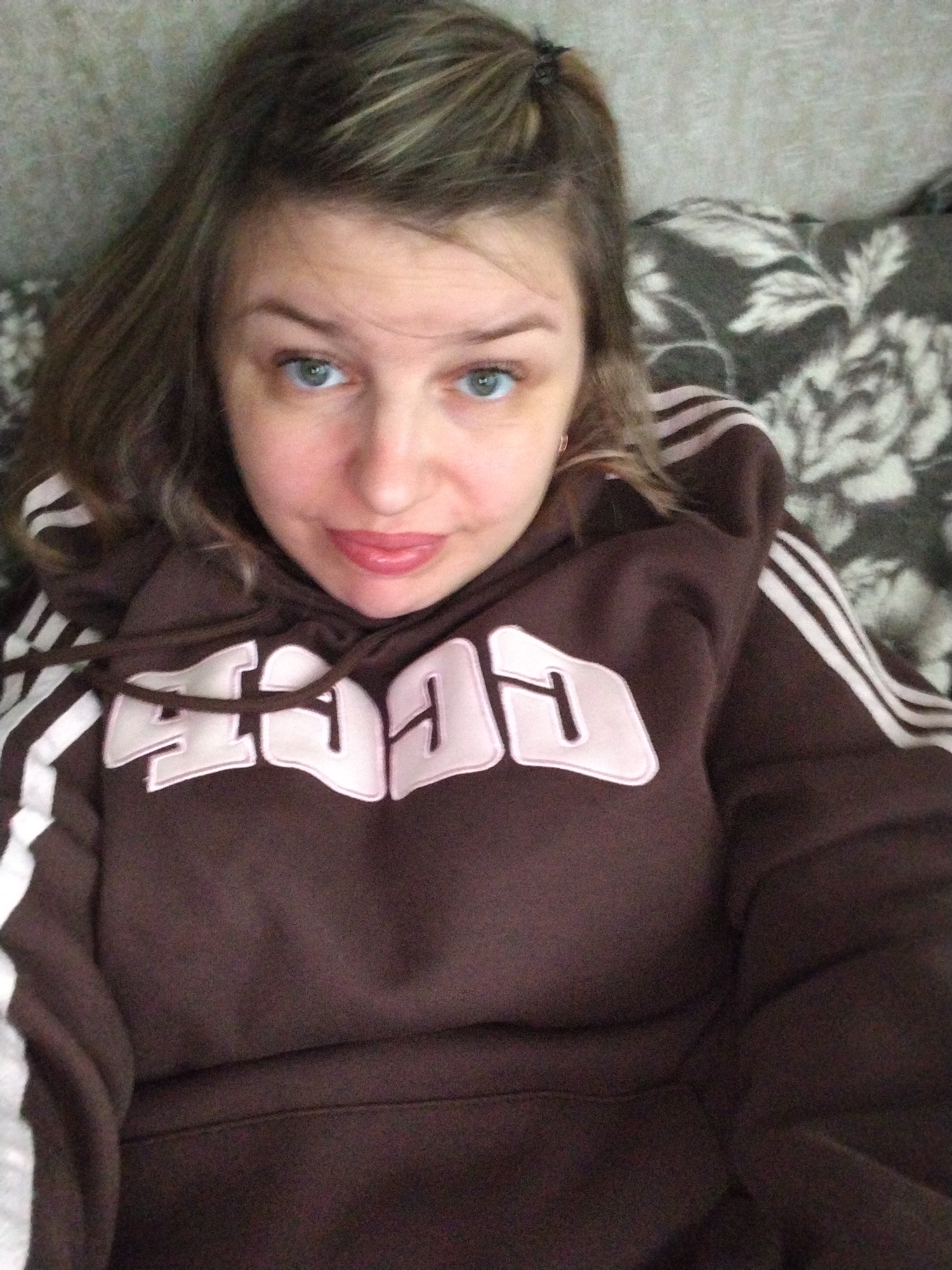 A woman is lying down, wearing a brown hoodie with white stripes on the sleeves and light pink lettering on the front, against a background of gray floral patterned fabric. She has light brown hair, blue eyes, and fair skin. Her lips are slightly pursed. The photograph is a close-up, and no specific location in the city of Yaroslavl, Russia, is identifiable.