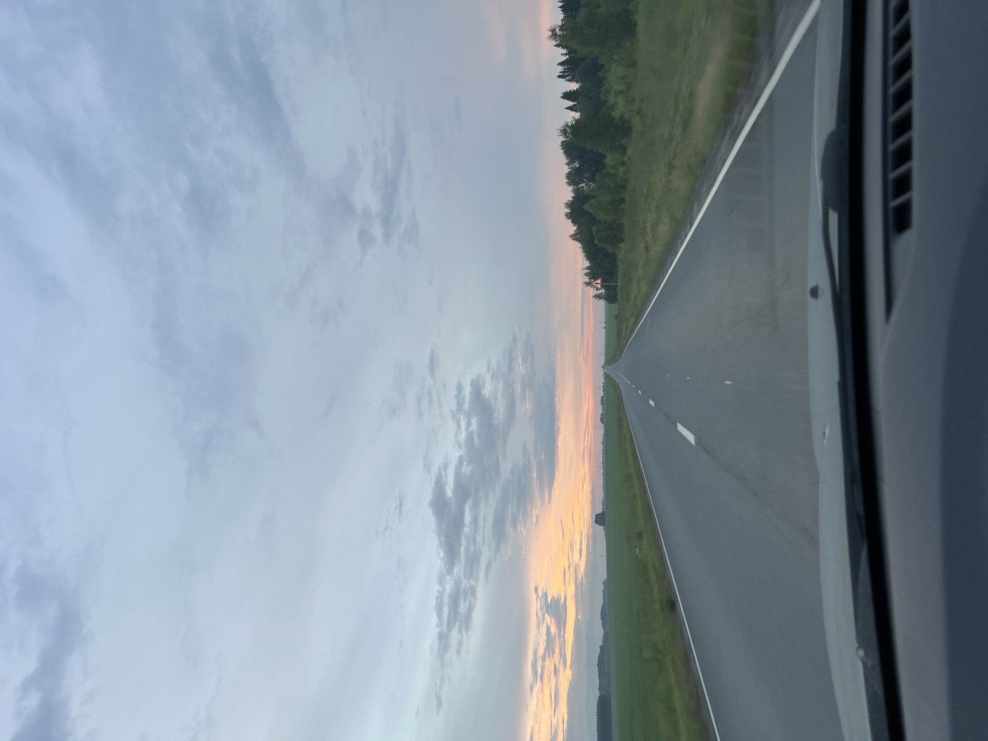 This media file captures a view from a vehicle traveling along a road in Поварёнки, Russia. The scene is dominated by a vast sky, displaying soft pastel colors of a sunset or sunrise, transitioning from pale blue to hues of pink and orange near the horizon. Clouds are scattered across the sky, adding texture to the atmospheric backdrop.
The road itself is paved and appears to be a straight, two-lane highway. White lines mark the edges and center of the road, indicating it is a well-maintained route. To the left of the road, a lush green field stretches out, bordered by a dense line of trees that form a dark silhouette against the colorful sky. To the right, the side of the vehicle is visible, including a windshield wiper and part of the car's interior trim. A road sign, a yellow triangle with a black symbol, is present near the treeline on the left side of the road.
The time of day is clearly either dawn or dusk, given the sky's coloration and soft light. The weather appears calm, with clear skies and no indication of rain or strong winds. The overall mood conveyed is one of peaceful travel and the serene beauty of nature during twilight. The perspective suggests the viewer is a passenger or driver, looking out through the windshield. There is no discernible text within the image.