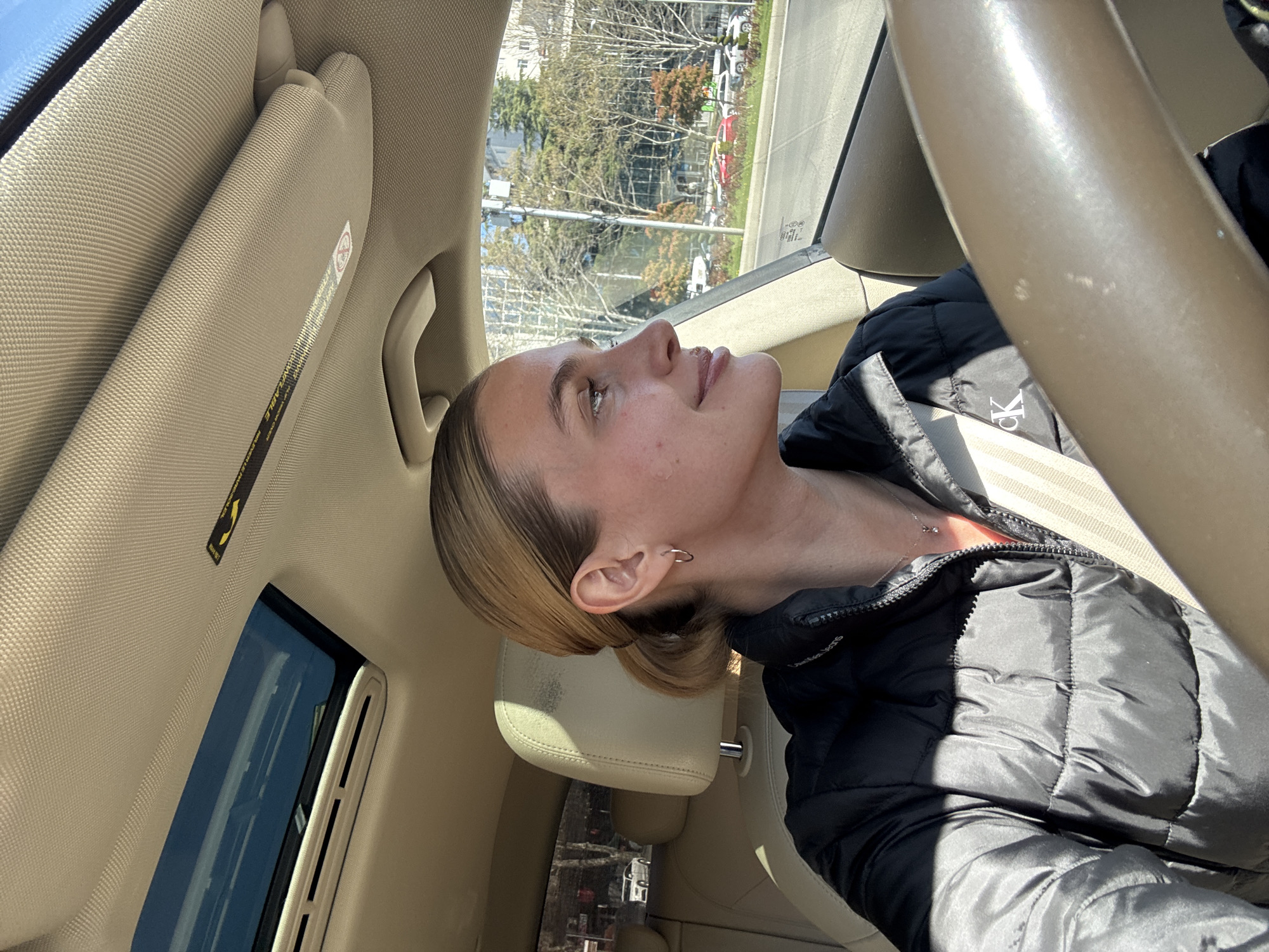 The image shows a woman in the driver's seat of a car. She is wearing a black puffer jacket and a seatbelt. Her hair is blonde and pulled back. A silver hoop earring is visible in her left ear. The interior of the car is light beige. Through the car window, buildings and trees are visible. Based on the architecture and vegetation, the location is possibly in the city of Tbilisi, Georgia, although there is no definitive evidence to confirm this. The driver is looking upwards.