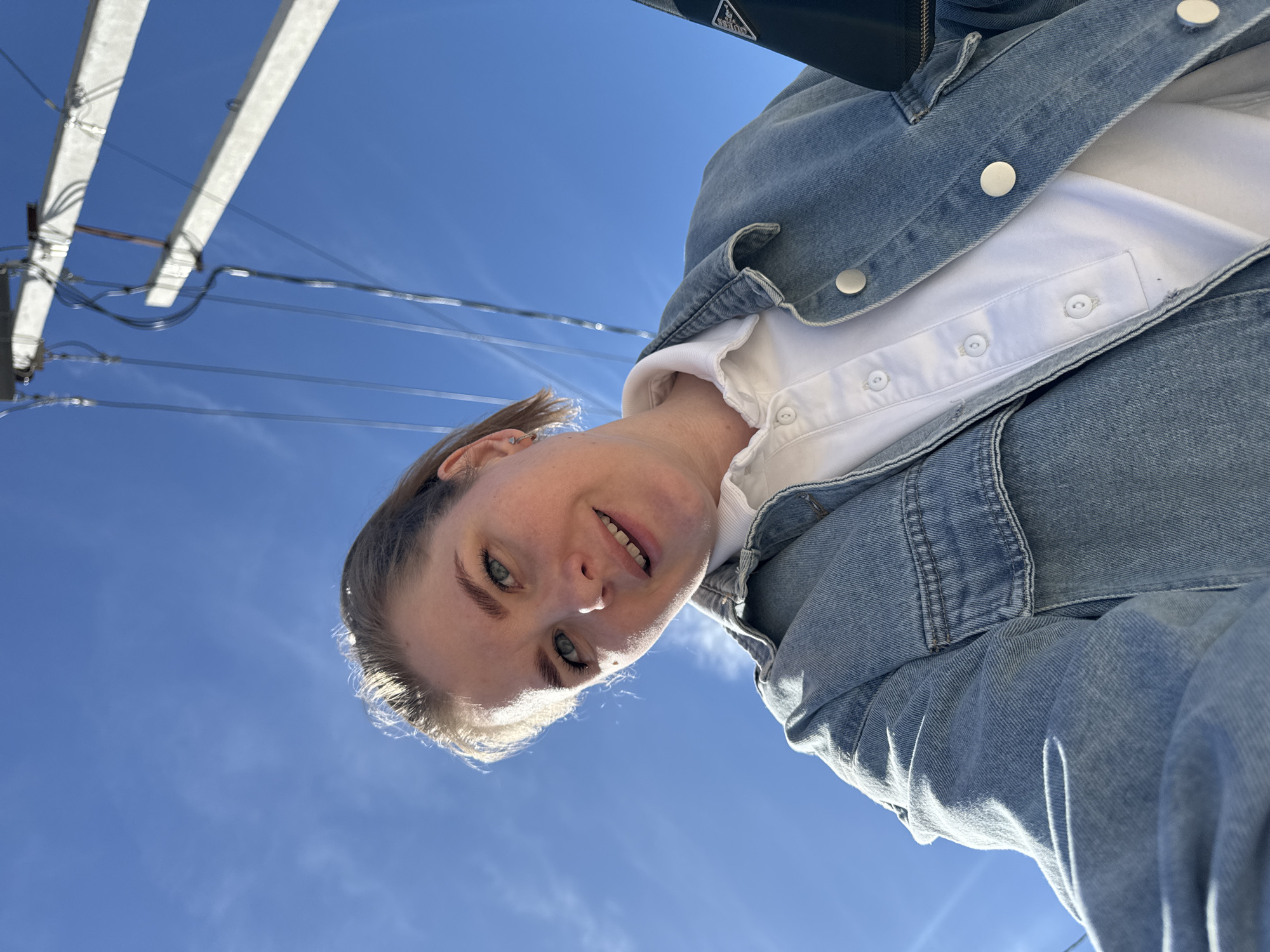 A low-angle medium shot shows a person wearing a light blue denim jacket and a white collared shirt against a clear blue sky. In the upper left, utility poles and power lines are visible. The person appears to be outdoors in the city of Ферма, Russia, based on the surrounding environment.