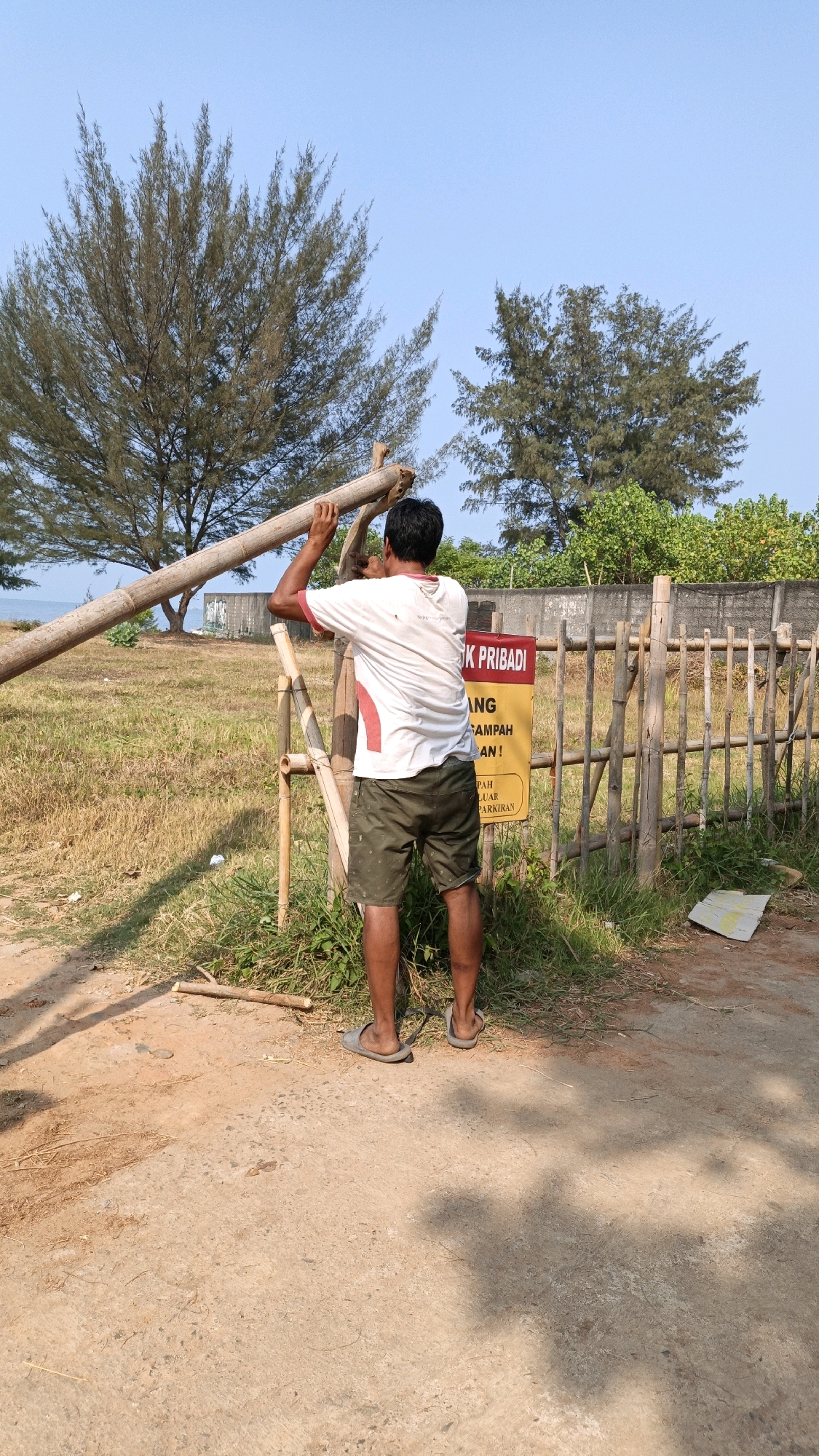 A man is standing in front of a bamboo fence, holding a bamboo pole. He is wearing a white t-shirt with red trim and olive green shorts. The fence is made of bamboo poles and is approximately 5 feet high. Behind the fence, there is a concrete wall and lush green vegetation. A yellow sign with red lettering is attached to the fence. The ground in front of the fence is dirt and gravel. The sky is clear and blue. The scene appears to be in Cikal, Indonesia, possibly near a beach or coastal area, given the vegetation and light.