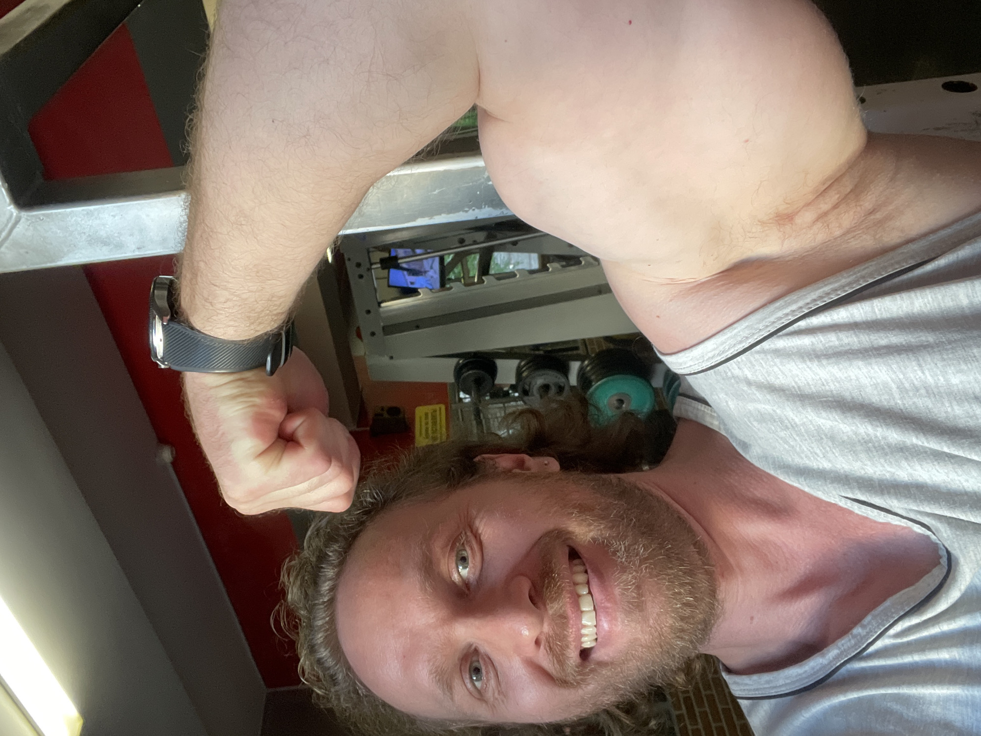 A fair-skinned man with curly, ginger hair and a beard smiles at the camera while flexing his bicep in a gym. He is wearing a light grey t-shirt with dark trim around the neckline and armholes. A black smartwatch is visible on his left wrist. Behind him, a weight rack with various weights and a television screen can be seen. The background suggests an indoor gym environment. The man is positioned in the foreground, with his face and bicep prominently featured. The lighting appears to be artificial, possibly from overhead gym lights, casting a warm glow on the scene. The image captures a moment of personal achievement or display of fitness within a gym setting. There is no identifiable indication of the location being Saint Petersburg, Russia.