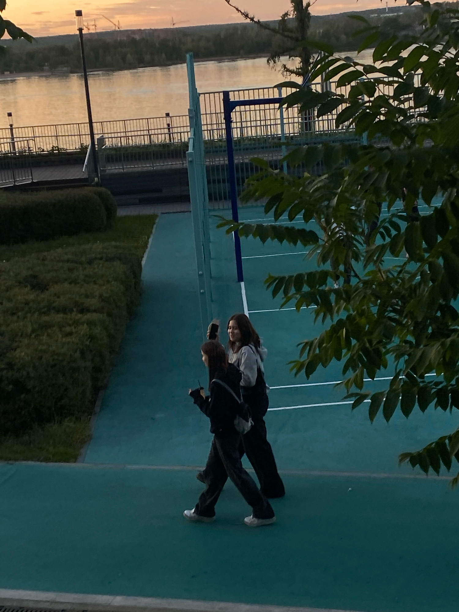 The image shows two young women walking on a teal-colored outdoor sports court. The woman in the foreground is wearing a black hoodie and dark pants, and is holding a drink with a straw. The woman behind her is wearing a gray hoodie and dark pants, and is holding a cell phone. They are walking away from the viewer, towards the right side of the frame. In the background, there is a body of water, likely the Kama River, with a treeline and a hazy sky. A fence separates the sports court from a walkway and a grassy area. The scene appears to be set in the city of Perm, Russia, possibly near the embankment. The time of day seems to be late afternoon or early evening, given the soft lighting and warm tones of the sky.