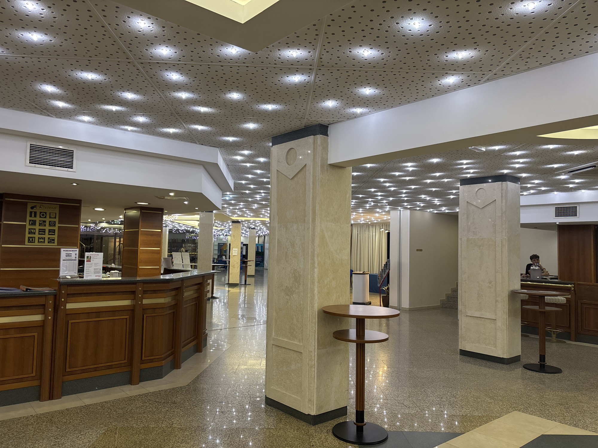 The image depicts the interior of a hotel lobby in Varna, Bulgaria, featuring a polished marble floor reflecting numerous small, bright lights from the perforated ceiling panels above. In the foreground, a light brown, multi-tiered circular side table with a dark base stands next to a large, beige stone pillar. To the left, a long, dark wood reception desk with gold accents is visible, with signs and informational pamphlets displayed behind it. Further back, another similar side table is positioned. In the background, a staircase ascends to the right. A person is seated at a counter on the far right, looking at a laptop. The overall ambiance is brightly lit and modern, suggesting a well-maintained hotel environment.