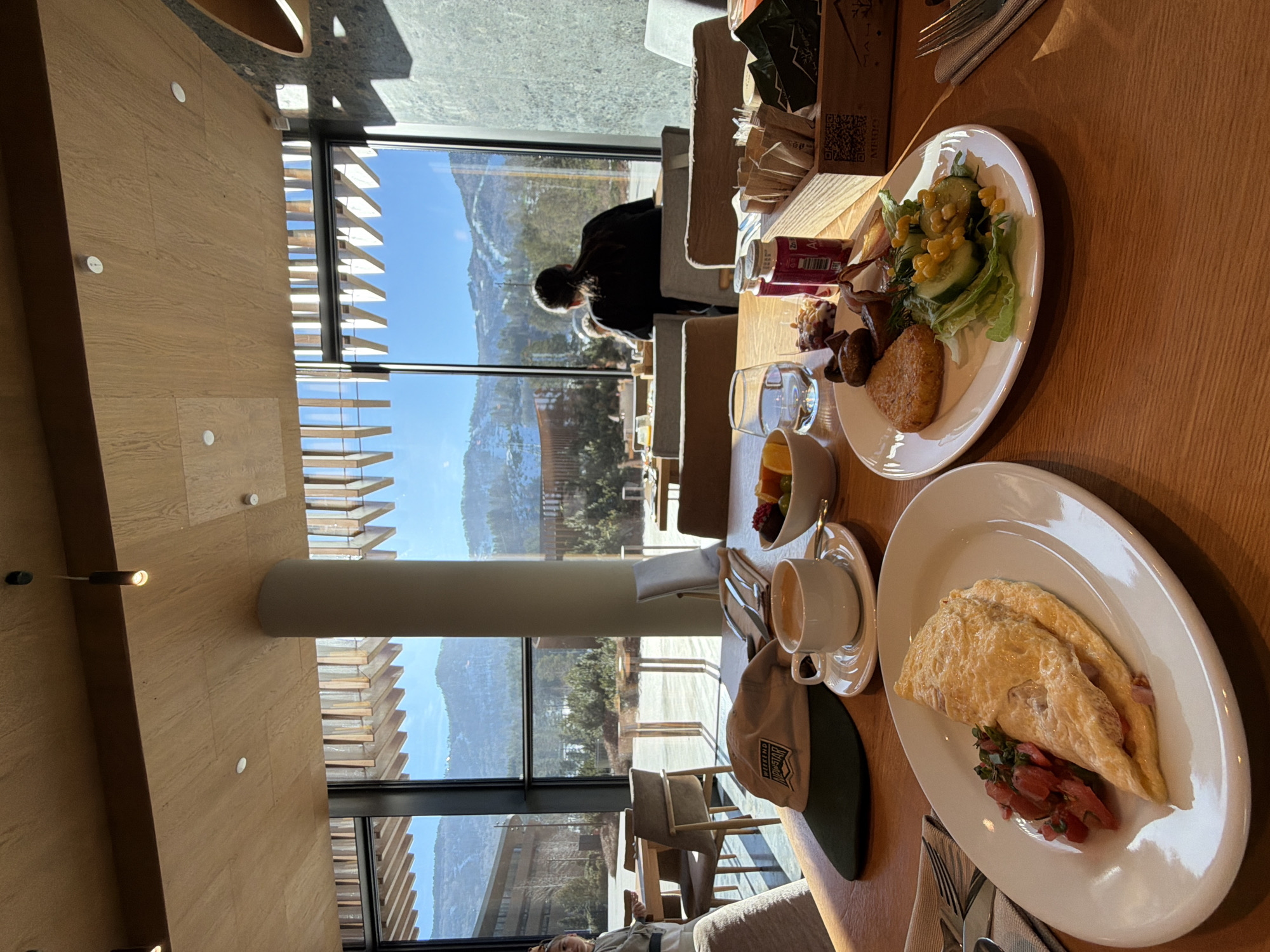 The image shows a breakfast setting with a view of mountains, possibly in or near Озёрное, Russia. Two plates of food are visible: one with an omelet and some chopped fruit, and another with a salad, hash brown, and some dark meat. A cup of coffee and a small bowl of fruit are also on the table, along with a hat. Through the large windows, a mountainous landscape with trees and some structures is visible under a blue sky. Another person can be seen sitting at a table further away by the window.