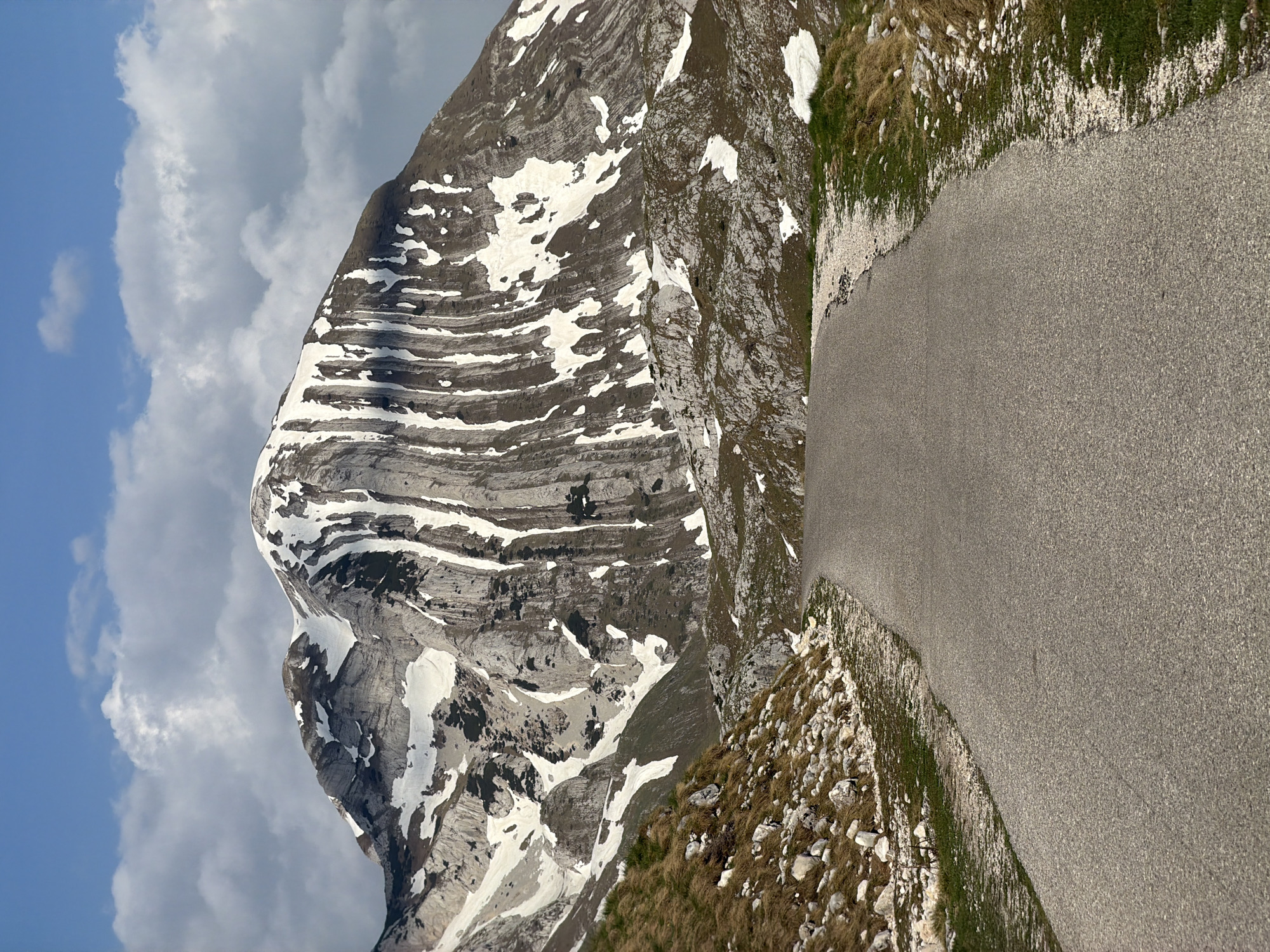 The image depicts a mountainous landscape in Boričje, Montenegro. A large, layered rock mountain occupies the left side of the frame, partially covered with snow patches. The sky above is a mix of blue and cloudy patches. A paved road is visible on the right side of the image, with grassy terrain and rocky patches bordering it. There are no people visible in the scene. The primary focus is on the mountain and the surrounding natural landscape.