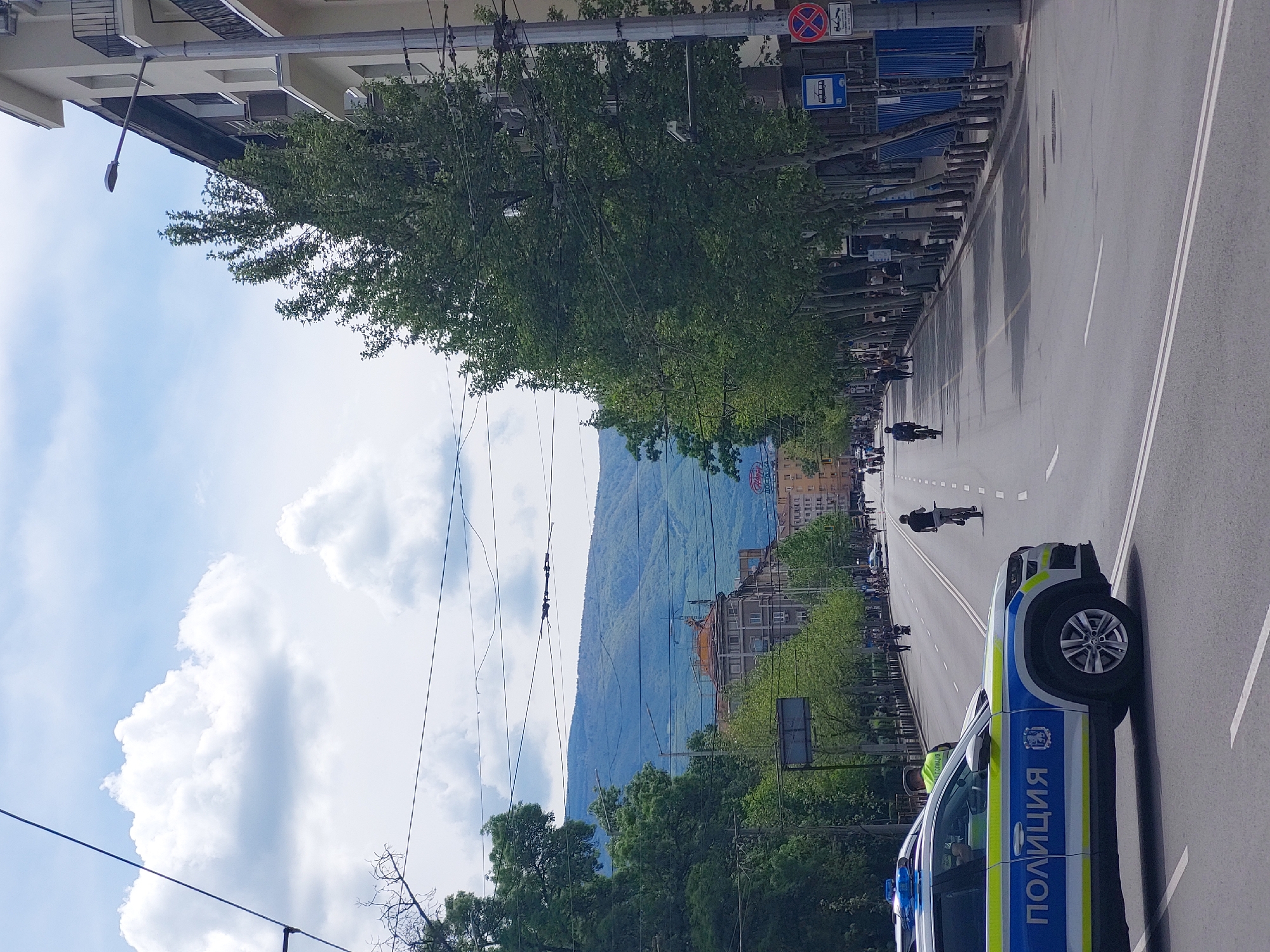 The image shows a street in Sofia, Bulgaria, with a police car on the right side of the frame. The word "ПОЛИЦИЯ" (Police) is prominently displayed on the car. The street is wide with multiple lanes and white lane markings. In the distance, there is a view of Vitosha Mountain. A line of people are walking and bicycling down the center of the road, suggesting a organized event or procession. The street is lined with trees and buildings. There is a street sign and bus stop on the left side of the street. There is construction on the side walk of the street. The sky is partly cloudy.