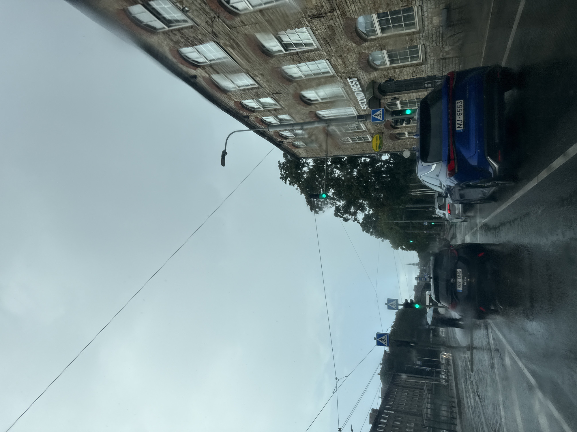 The image depicts a rainy street scene in Tallinn, Estonia. The view is from a vehicle driving on a wet road. In the foreground, a dark-colored car is visible, followed by a blue Lexus with license plate NU-6553. Beyond these vehicles, traffic lights are visible, one of which is green. Pedestrian crossing signs are posted on the sidewalks.
To the right of the road, a multi-story brick building with arched windows and the signage "SECOND BEST" is prominent. The building appears to be old, with visible brickwork. Trees line the street in front of and beside the building.
On the left side of the road, there are buildings and what appears to be a tram line with overhead wires. Two pedestrians with umbrellas are walking in the distance, one of them holding a white umbrella. The sky is overcast and the overall atmosphere suggests a gloomy, wet day. The street is reflective due to the rain, indicating significant moisture on the road surface.
