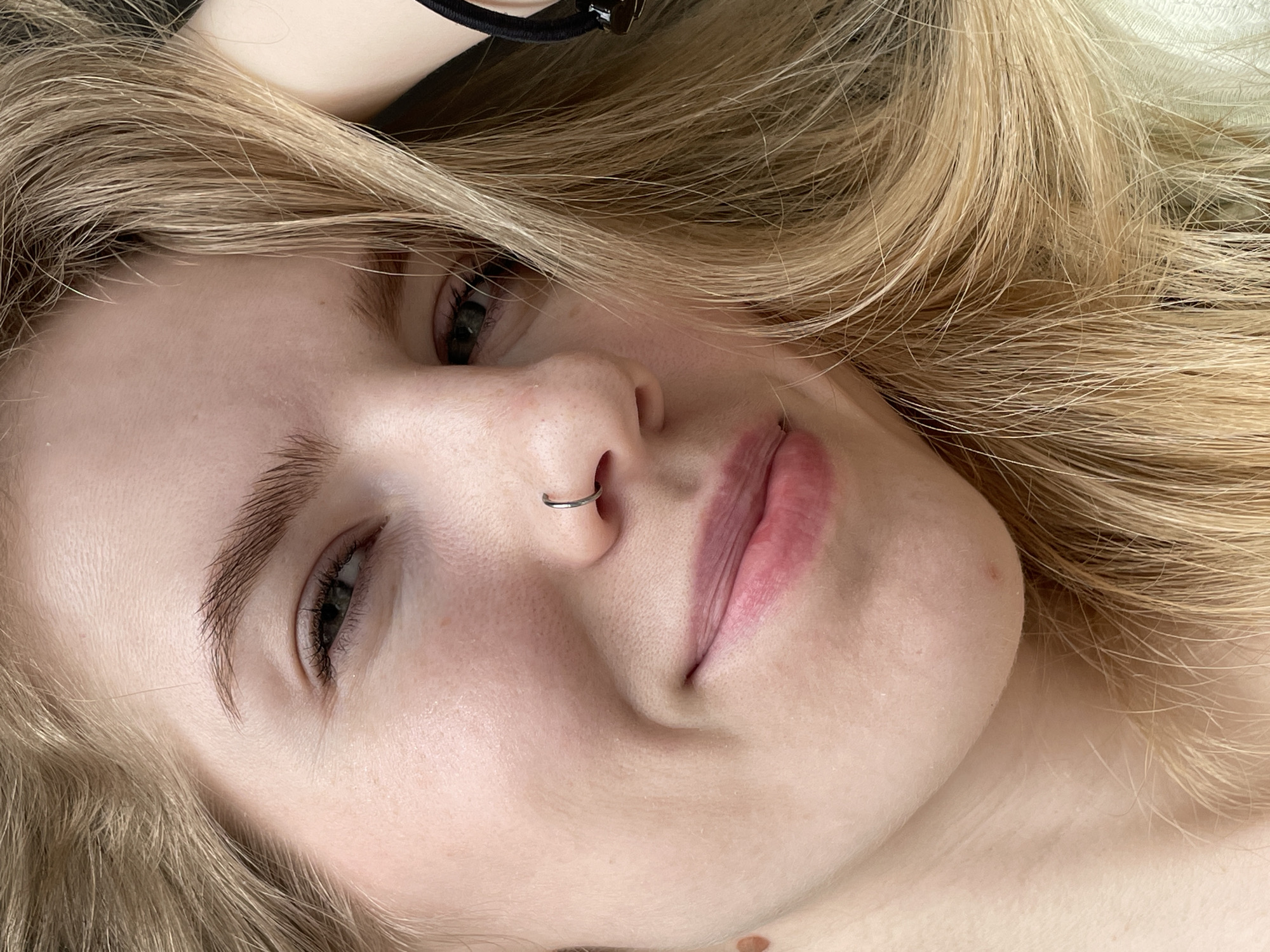 A close-up, high-angle portrait features a young woman with fair skin and long, wavy blonde hair, presented in a casual, relaxed pose. She appears to be lying down, looking directly at the camera with a subtle, relaxed expression that could be interpreted as a slight smile or content smirk. Her eyes are open, and her natural eyebrows are visible.
A notable detail is a silver-colored septum piercing (nose ring) adorning her nose. Her lips are slightly parted, revealing their natural pink hue. On her chin and upper neck area, a few small moles or freckles are visible. To the upper left of the frame, part of a black hair tie or scrunchie is seen on what appears to be her arm or wrist, blending into the blurred background. Her blonde hair frames her face, with strands falling across her forehead and around her head. The lighting is soft and even, suggesting an indoor setting with diffused light. The background is largely out of focus, showing indistinct light tones that could be fabric or a wall. There is no visible text in the image. This scene takes place in Izhevsk, Russia.