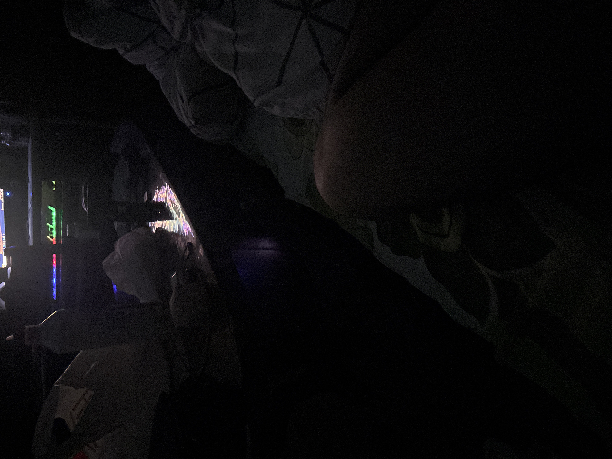 The photograph, likely taken indoors, presents a scene in near-total darkness. The top of the frame is dominated by what appears to be bedding, possibly a blanket or pillows, with geometric patterns visible in the dim light. Below this, a large dark form obscures much of the scene. In the lower-left quadrant of the image, illuminated by multi-colored lights, there is a desk with a computer. Various objects are scattered around the desk. The location in Yerevan, Armenia cannot be confirmed based on visual evidence.