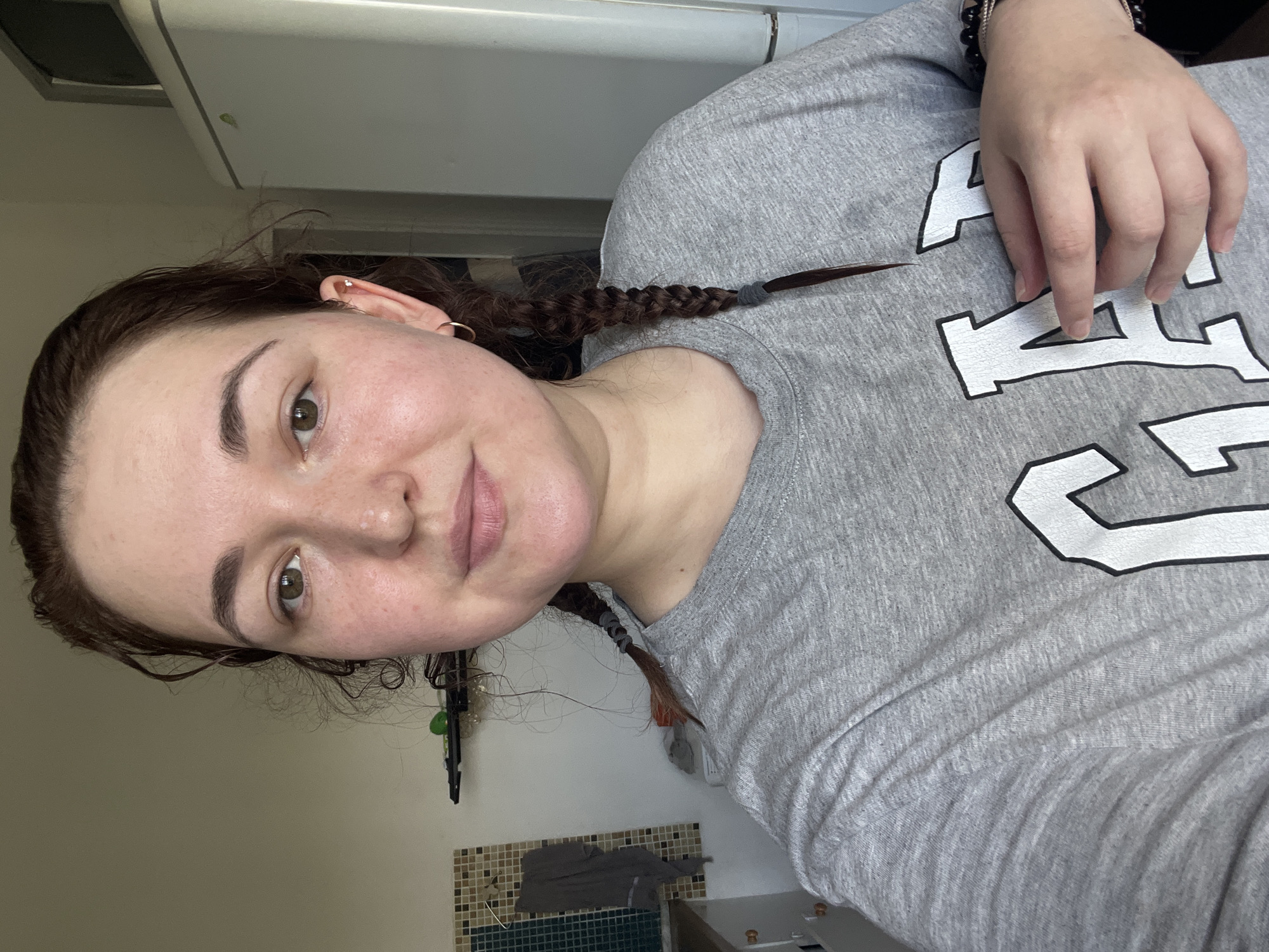 The image shows a young woman with brown hair braided into two pigtails, wearing a gray t-shirt with the "GAP" logo. She has light skin with freckles and appears to be indoors, in what looks like a kitchen. In the background, a refrigerator and kitchen cabinets are visible. The scene appears to be domestic. The image does not contain any information that would suggest a specific location in Chelyabinsk, Russia.