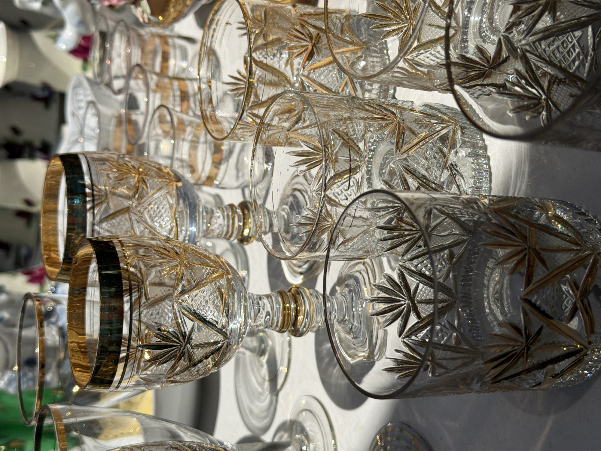 A close-up, eye-level, outdoor shot captures an assortment of clear crystal glasses with gold trim and etched starburst patterns arranged on a white surface. The lighting suggests it is daytime, with strong shadows cast by the glasses. The background is blurred, showing hints of other decorative items, possibly in a market or shop setting. There are no people or animals visible, and no obvious actions are taking place. The scene is static and focuses on the arrangement and detail of the glassware.