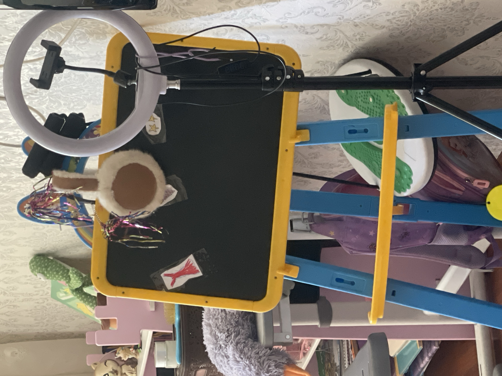 The image shows a children's art easel with a blackboard in the center. The easel is blue and yellow. There are several items attached to the blackboard, including a drawing of a red dress and two yellow stars. A ring light with a phone holder is positioned in front of the blackboard. Behind the easel, there are various toys and clutter, including a plush green dinosaur toy, a pink toy castle, and a fuzzy purple toy. The background consists of patterned wallpaper. The scene appears to be indoors, likely in a child's room or play area.