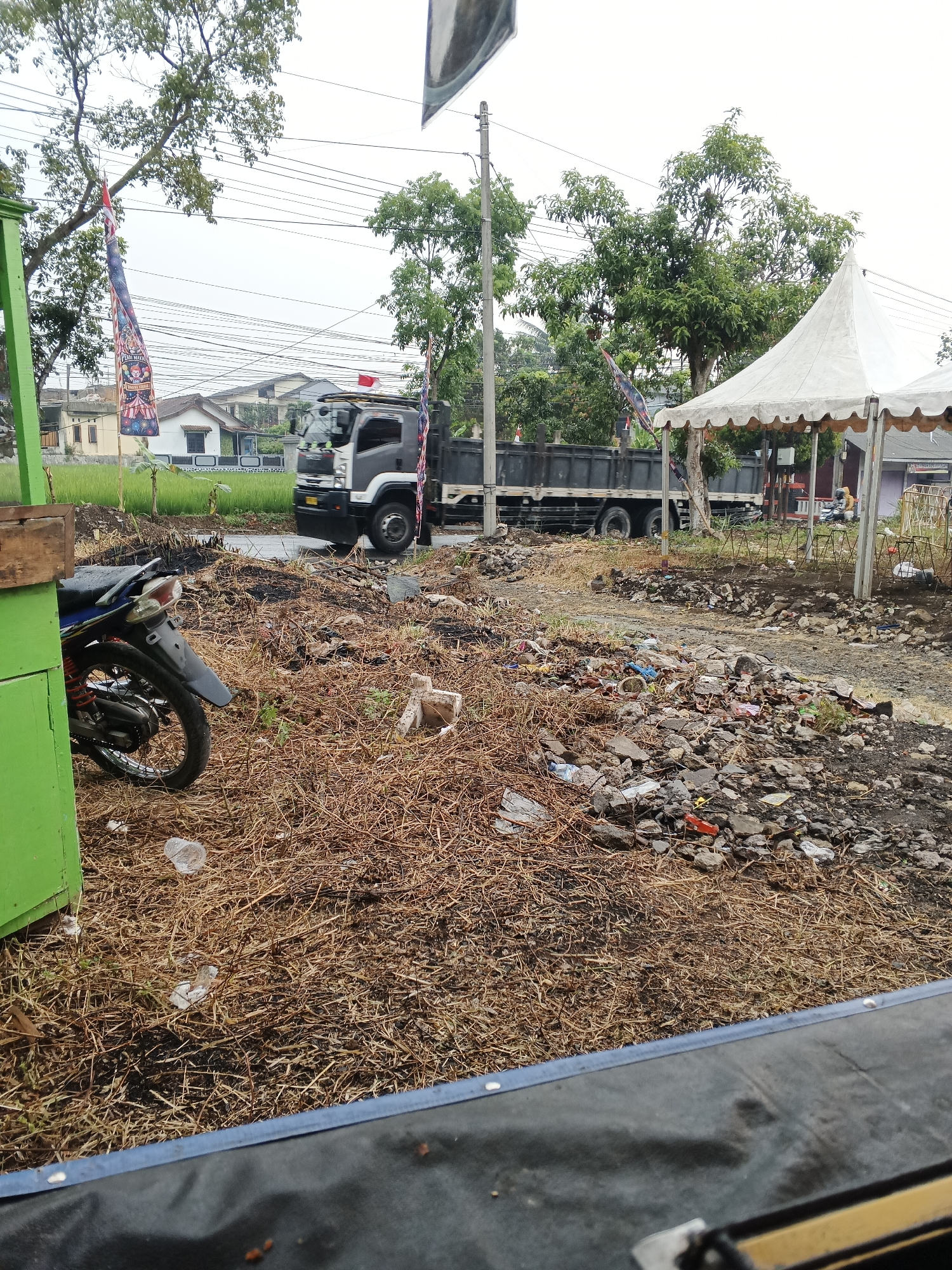 The image depicts an outdoor scene with a large amount of debris and dry vegetation covering the ground. In the foreground, a portion of a green structure is visible on the left, with a motorcycle parked next to it. Further into the scene, a large gray dump truck is driving on a road. A white tent is set up on the right side of the image. Various flags are displayed, including one with colorful designs and another that appears to be a national flag. Buildings and trees are present in the background, suggesting an urban or semi-urban environment. The presence of a dump truck, a tent, and a road indicates a possibly industrial or event-related setting, or simply a location undergoing some form of activity or cleanup. The scene appears to be located in Sukabumi, Indonesia, given the context provided.