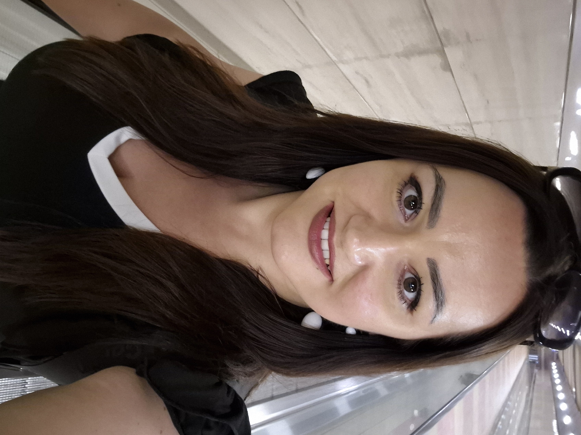A medium shot depicts a woman posing for a selfie or photograph indoors. She has long, brown hair and fair skin, wearing a black top with a white trim along the neckline. Her eyes are brown, and she is smiling, showing her teeth. Sunglasses are perched on the side of her head. The background appears to be a modern public space or transportation hub, possibly an airport or mall.