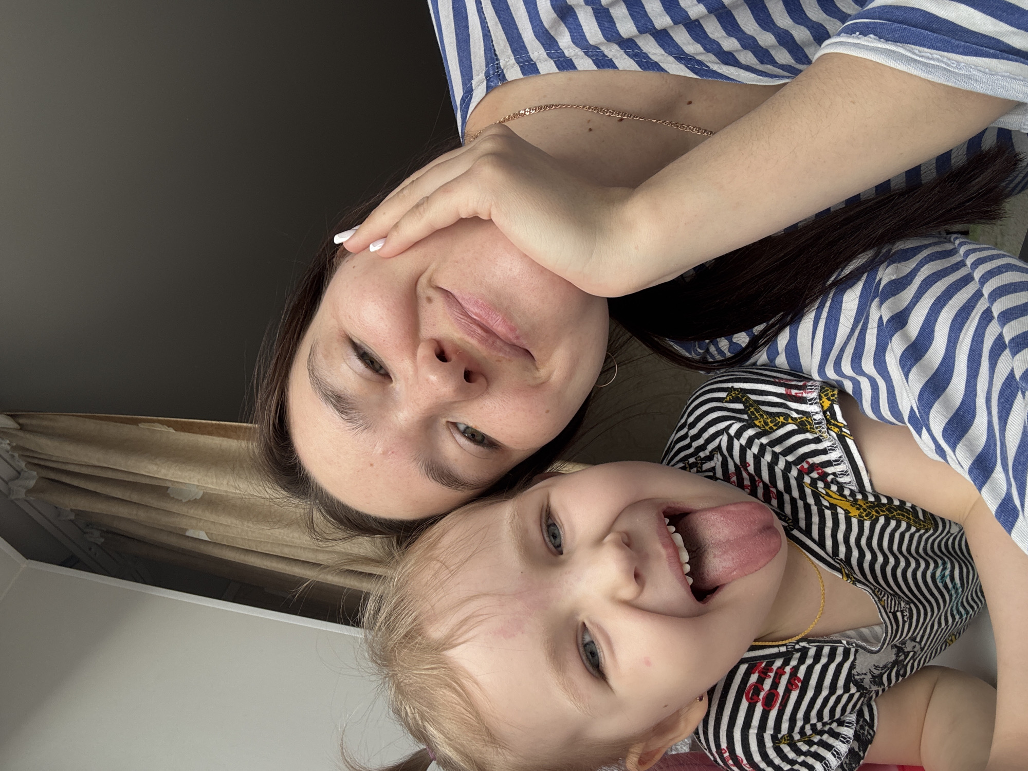 A selfie taken indoors, possibly in an apartment. A woman with dark hair and a blue-and-white striped shirt is holding her hand near her face. Next to her is a young girl with blonde hair in pigtails, wearing a black-and-white striped shirt with a gold necklace, sticking her tongue out. The background shows a brown curtain, a white wall, and a dark grey wall or surface. The location is likely Chaikovsky, Russia, though no identifying landmarks are visible.