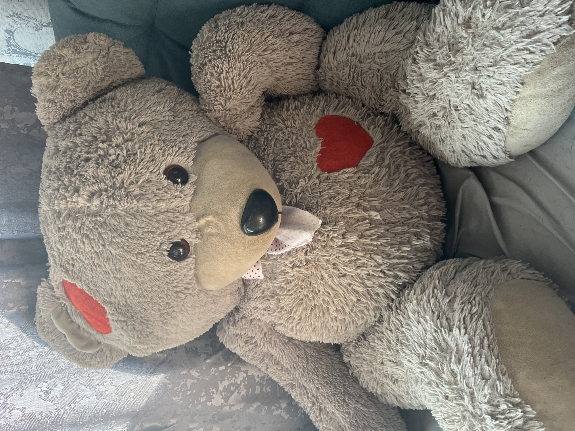 A large, fluffy, light brown teddy bear is seated on a plush chair. The bear has dark brown button eyes, a black nose, and a light brown muzzle. It wears a white bow with red polka dots around its neck. A red heart patch is visible on its chest, and another red patch is on its left ear. The bear is positioned in a relaxed, seated pose, with its arms resting on its lap. The background consists of a textured, light-colored curtain to the left and a dark teal chair. The overall setting appears to be a domestic indoor environment. The image was taken in Verkhnedneprovskiy, Russia.