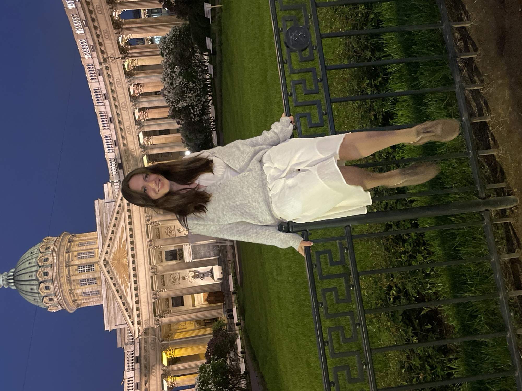 A nighttime shot presents a woman leaning against a dark-colored metal fence. She is wearing a white dress and a grey sweater. Her long, dark hair is cascading down her shoulders. Behind her is a green lawn. In the background, illuminated by artificial light against a dark blue sky, is the architectural edifice of Kazan Cathedral in Saint Petersburg, Russia.