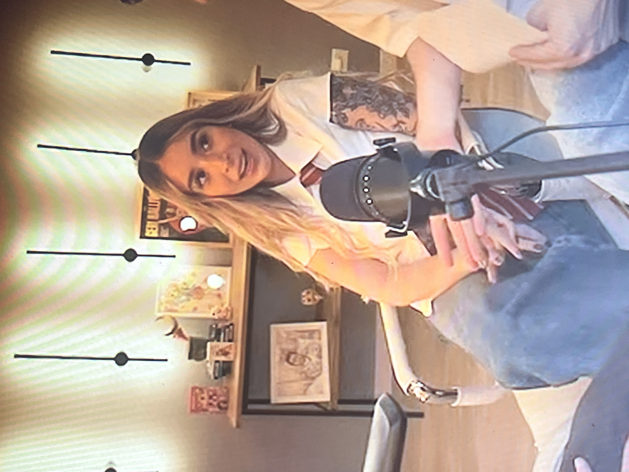 The image depicts a young woman seated in a chair, facing forward and speaking into a microphone. She has long, blonde hair and is wearing a white collared shirt with a red and striped tie, resembling a school uniform. She has tattoos on her right arm. The setting appears to be indoors, possibly a studio or room, with a shelf behind her displaying various items, including framed pictures and decorative objects. There are also vertical lines on the wall behind the shelf, which might be decorative elements or part of a lighting fixture. The overall lighting suggests it is likely daytime or well-lit indoors. There is no visual information in the image that allows for the identification of the specific location within the city of Santa Ana, Venezuela.