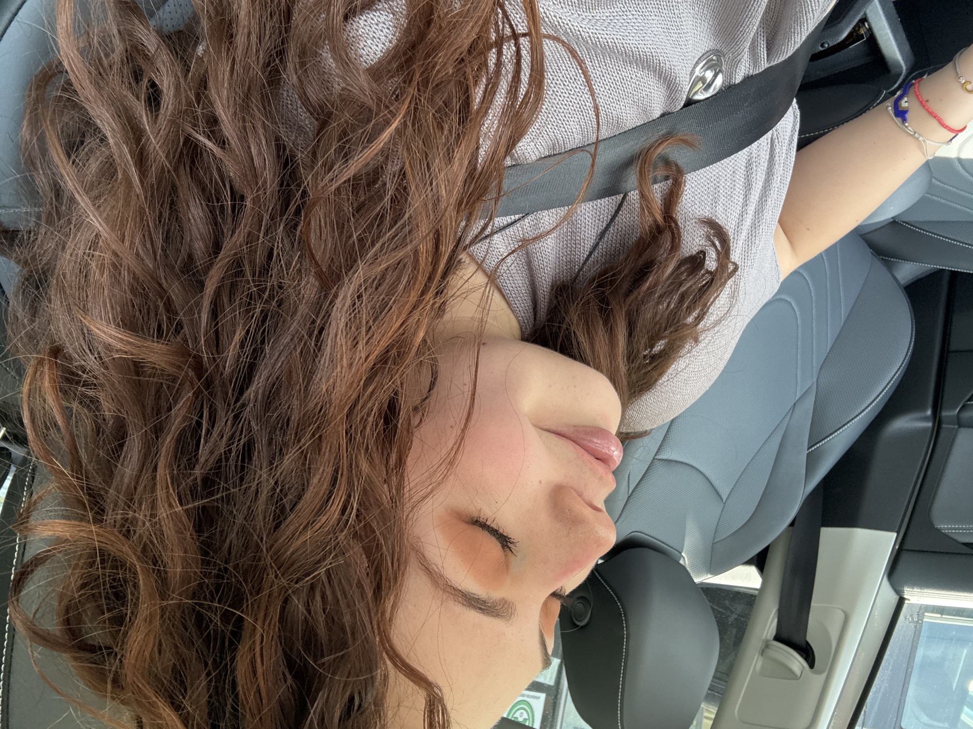 A woman with long brown curly hair is in a car, wearing a gray sweater and a seatbelt. She is likely in Mexico City, Mexico based on the Spanish-language sticker visible in the window near her face. She is likely a passenger in the vehicle. She is wearing a light makeup around her eyes and several bracelets on her right wrist. The interior of the car has gray leather seats.