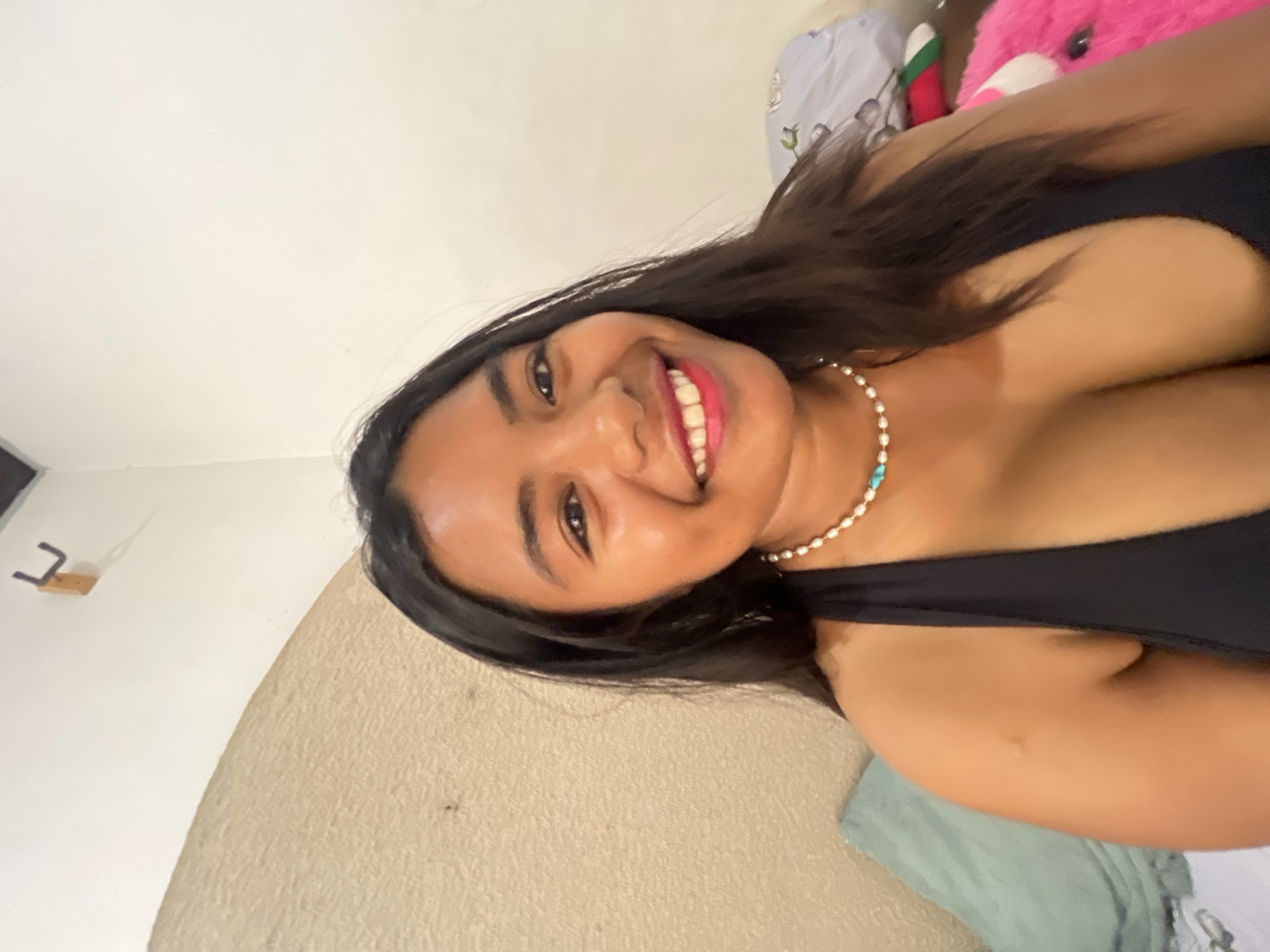 A young woman with dark hair and a bright smile is posing for a selfie. She is wearing a black halter top and a pearl and turquoise necklace. Her skin is bronzed, and she has a light dusting of freckles across her nose and cheeks. She is seated in a room with a light-colored, textured wall behind her. To her left, a teal pillow is visible. To her right, a pink stuffed animal and some patterned fabric are in the background. A wooden hook is mounted on the wall in the upper left portion of the frame. The lighting is bright and suggests an indoor setting. The image appears to have been taken in Denpasar, Indonesia.