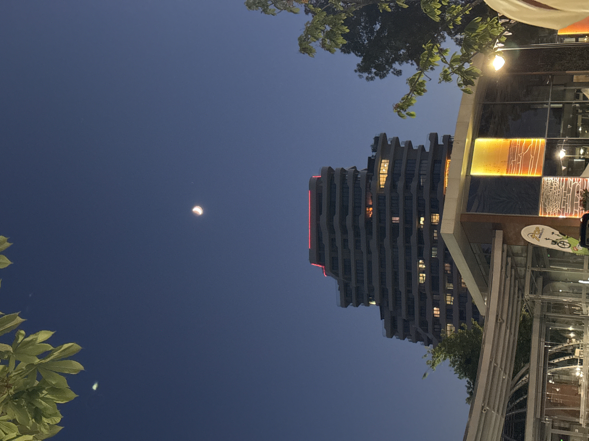 This is a nighttime photograph of a unique, tiered building in Burgas, Bulgaria. The building has a modern design with multiple balconies or setbacks, and red lighting accents its top edge. Numerous windows are illuminated, suggesting activity within. In the foreground, there is a glass structure, possibly a cafe or part of a shopping area, with artistic panels on its exterior that glow with warm orange and yellow light. Trees with green leaves are visible on the right and left sides of the image, framing the scene. A crescent moon hangs in the dark blue sky, high above the building, and a faint celestial object is visible near some leaves in the upper left quadrant. The overall impression is of a vibrant urban evening.