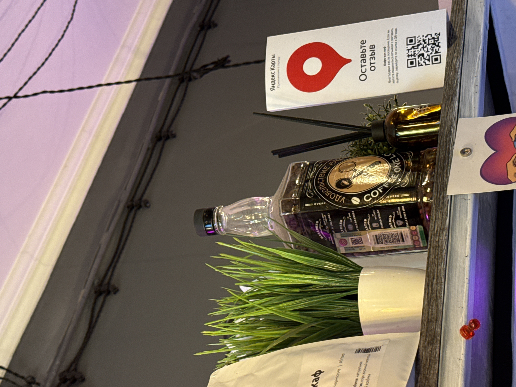 The image depicts a shelf in a dimly lit establishment, likely a cafe or shop, with various objects displayed. On the left, a white paper bag with Cyrillic text is partially visible, suggesting it contains coffee or a similar product. Next to it, a potted artificial green plant with long, thin leaves stands in a white pot. To the right of the plant is a distinctive brown glass bottle, recognizable as Jack Daniel's Tennessee Whiskey, with its iconic label and black cap. Further to the right, a small, amber-colored glass bottle with a diffuser stick is placed. Adjacent to this is a white card with Cyrillic text and a red location marker graphic, indicating "Yandex Maps" and an invitation to "Leave a review." A QR code is printed at the bottom of this card. The background shows a dark wall and a string of lights overhead. The shelf itself appears to be made of wood. Below the shelf, two small, red, circular objects, possibly berries or decorative elements, are visible against a purple light. The overall scene suggests a point of sale or display area within a commercial space. The presence of "Yandex Maps" and Cyrillic text indicates the location is in Russia, most likely Moscow, given Yandex's dominance in the Russian market and the prevalence of Cyrillic script.