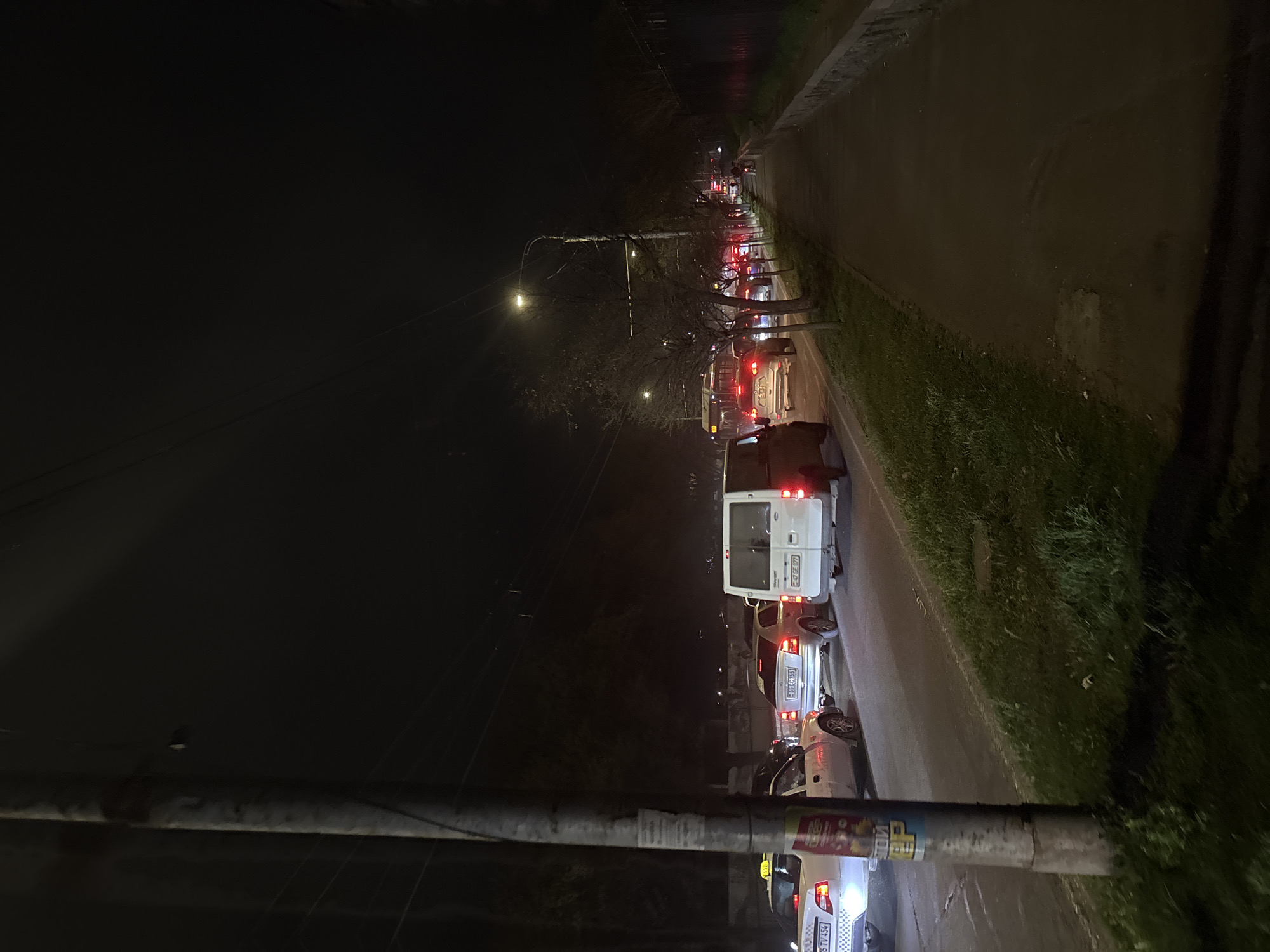 The image depicts a nighttime street scene in Yerevan, Armenia. Several vehicles are stopped on a road, suggesting a traffic jam. The vehicles include a white van with license plate "E92EZ977" and a white sedan with license plate "E550CE95". A taxi can also be seen on the right, with license plate "515TV454". Streetlights illuminate the scene, and trees are visible along the roadside. A sidewalk with grass borders the road. A concrete pole is on the right in the image's foreground.