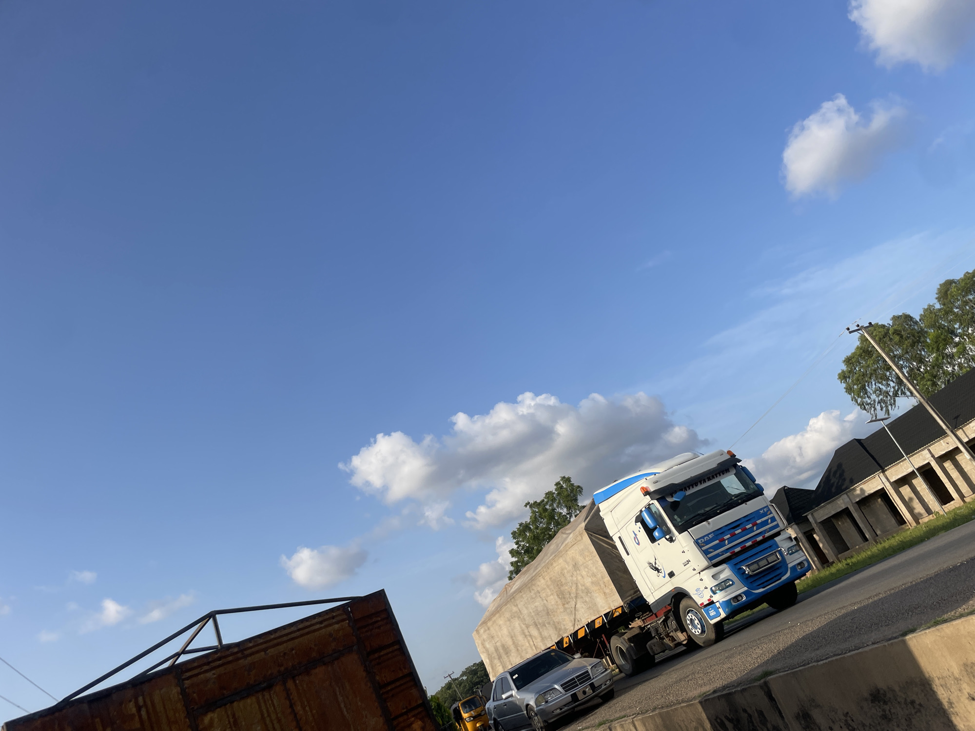 This media file depicts a street scene in Jalingo, Nigeria, under a clear blue sky with scattered white clouds. The sun appears to be low in the sky, suggesting either early morning or late afternoon, casting long shadows and illuminating the scene with warm light.
The primary subjects are a white and blue DAF XF truck pulling a large, tarp-covered trailer, and a silver sedan parked behind it. To the left, a section of a rusty metal structure is visible, possibly construction scaffolding or a discarded shipping container. In the background, a street runs alongside a row of buildings with dark, angled roofs, some of which appear to be under construction with exposed frameworks. Green trees line the street behind the buildings. A utility pole with wires is also present.
A smaller, yellow three-wheeled vehicle is partially visible behind the silver sedan, suggesting the presence of various modes of transportation. The overall scene conveys a sense of everyday activity on a road in Nigeria, with commercial and personal vehicles present.
Visible text on the truck reads "ATTUYA RATTYM" above the windshield and "DAF XF" on the front grille. The scene appears calm, with no overt indication of distress or urgency.