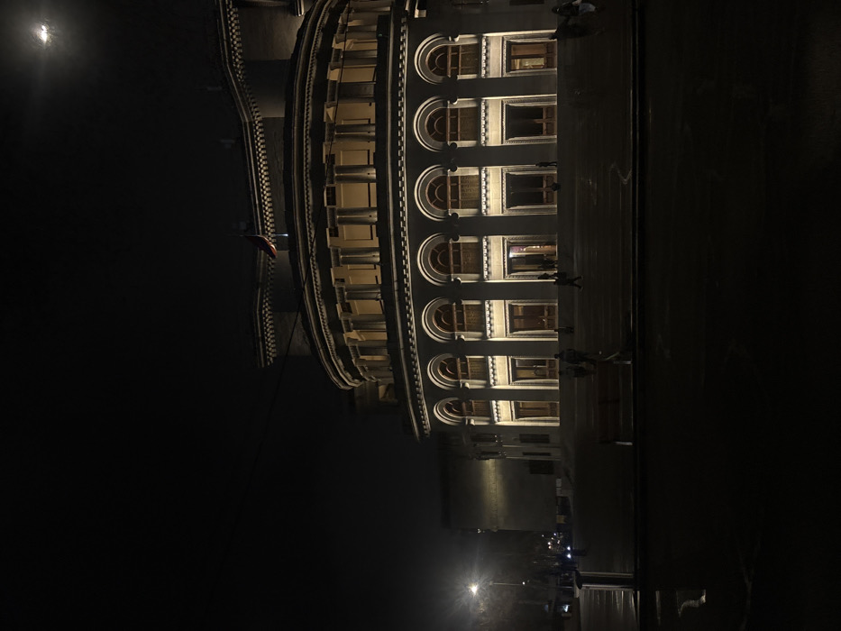The image depicts the Armenian National Academic Theatre of Opera and Ballet named after Alexander Spendiaryan in Yerevan, Armenia, at night. The prominent structure is illuminated by artificial warm-toned lights, highlighting its light-colored stone facade and neo-classical architectural features. The building's central rotunda section features two visible levels: a lower level with multiple arched doorways/windows separated by pilasters, and an upper level with rectangular windows set behind a colonnade. Intricate cornices and roofline details are discernible above. An Armenian tricolor flag is visible on the rooftop.
A wide, dark set of steps leads up to the building's entrances. Approximately 10-15 indistinct human figures, appearing as dark silhouettes, are present on these steps and near the building's lower entrances, suggesting pedestrian activity. In the lower-middle foreground, a dark-colored bench is visible on a dark surface, possibly pavement, which shows some reflective sheen, indicative of wetness, particularly on the left side where a strong street light illuminates a section. The street light is prominent on the far left, casting a bright glare. Beyond the street light, faint vehicle outlines and additional indistinct human figures are visible on a distant street. The sky is dark with minimal cloud cover, and a faint celestial body is discernible in the upper right quadrant. Trees are visible as dark masses in the background on the far left.