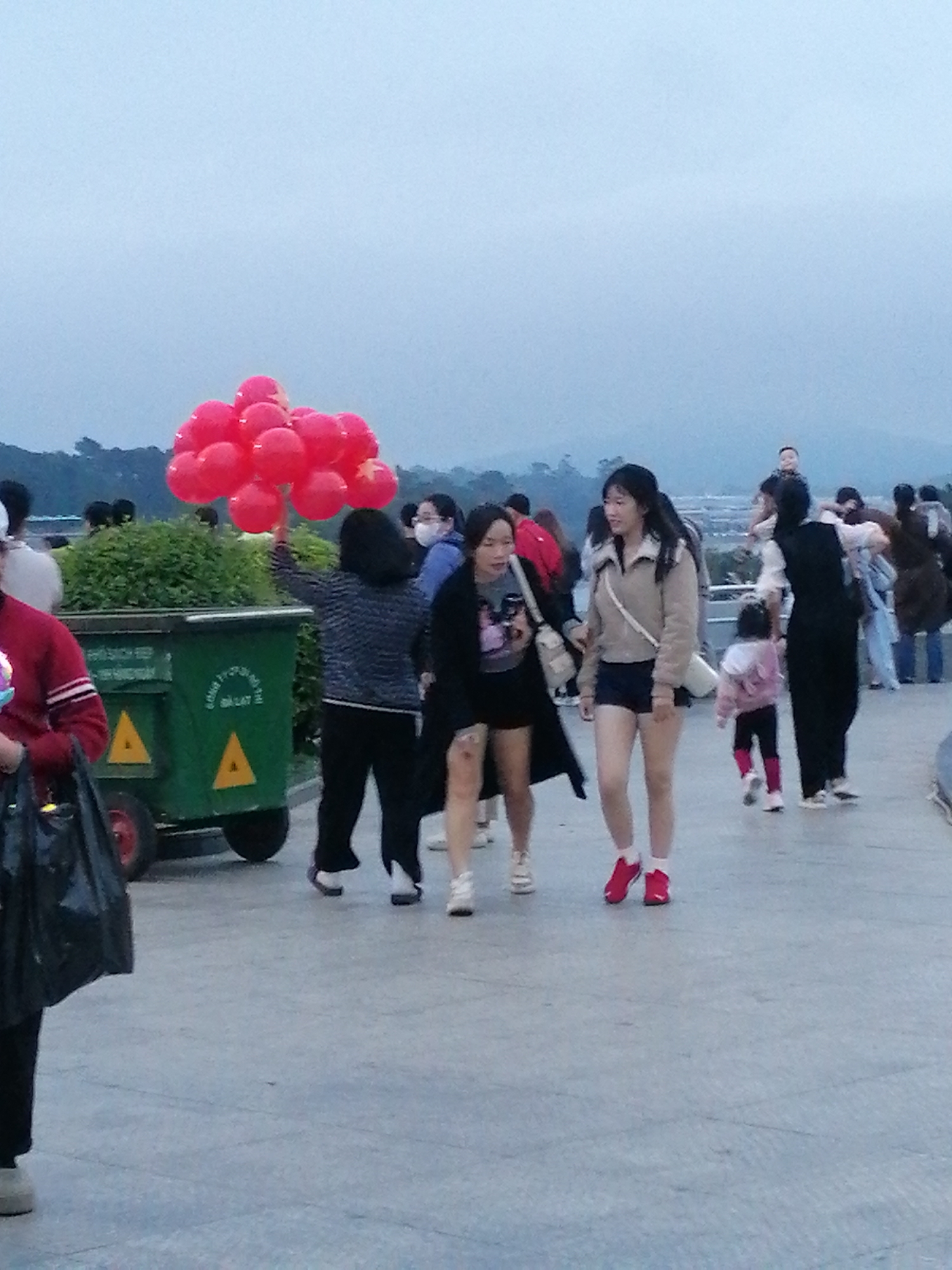 In Da Lat, Vietnam, a group of people are gathered in an outdoor setting near a green trash receptacle. A woman in the foreground is holding a large bunch of red balloons. To her right, another woman is walking away from the camera, wearing a beige jacket and dark shorts. Further in the background, several other individuals, including children, are visible, and some are walking or standing. The environment appears to be a paved walkway or plaza, with hints of greenery and a distant landscape under an overcast sky. The scene suggests a casual outing or public gathering.