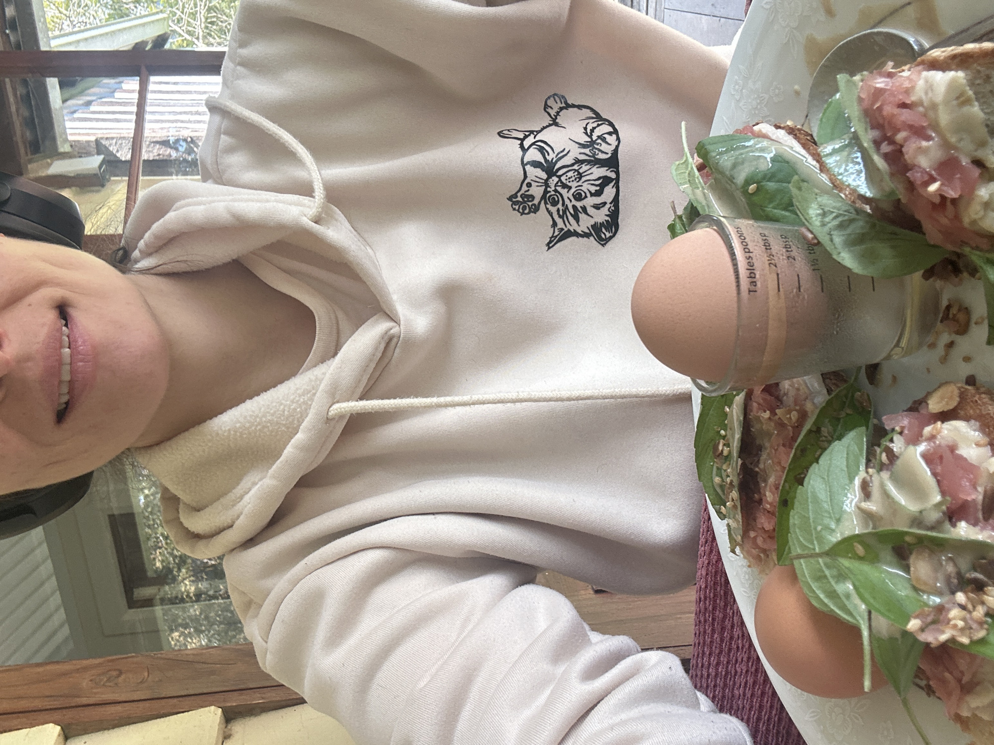 A person wearing headphones and a light-colored hoodie with a cartoon cat graphic is seated at a table outdoors, possibly on a deck or patio, as indicated by the wooden structures and foliage visible in the background. In front of them is a white plate with two whole brown eggs and what appears to be open-faced sandwiches topped with green leaves and a pinkish ingredient. A small glass measuring cup with liquid and markings for tablespoons is also on the plate, holding one of the eggs. The person's face is partially visible, showing a smile and their mouth. The environment suggests a casual meal in a natural setting, possibly in Thora, Australia, given the vegetation and outdoor ambiance.