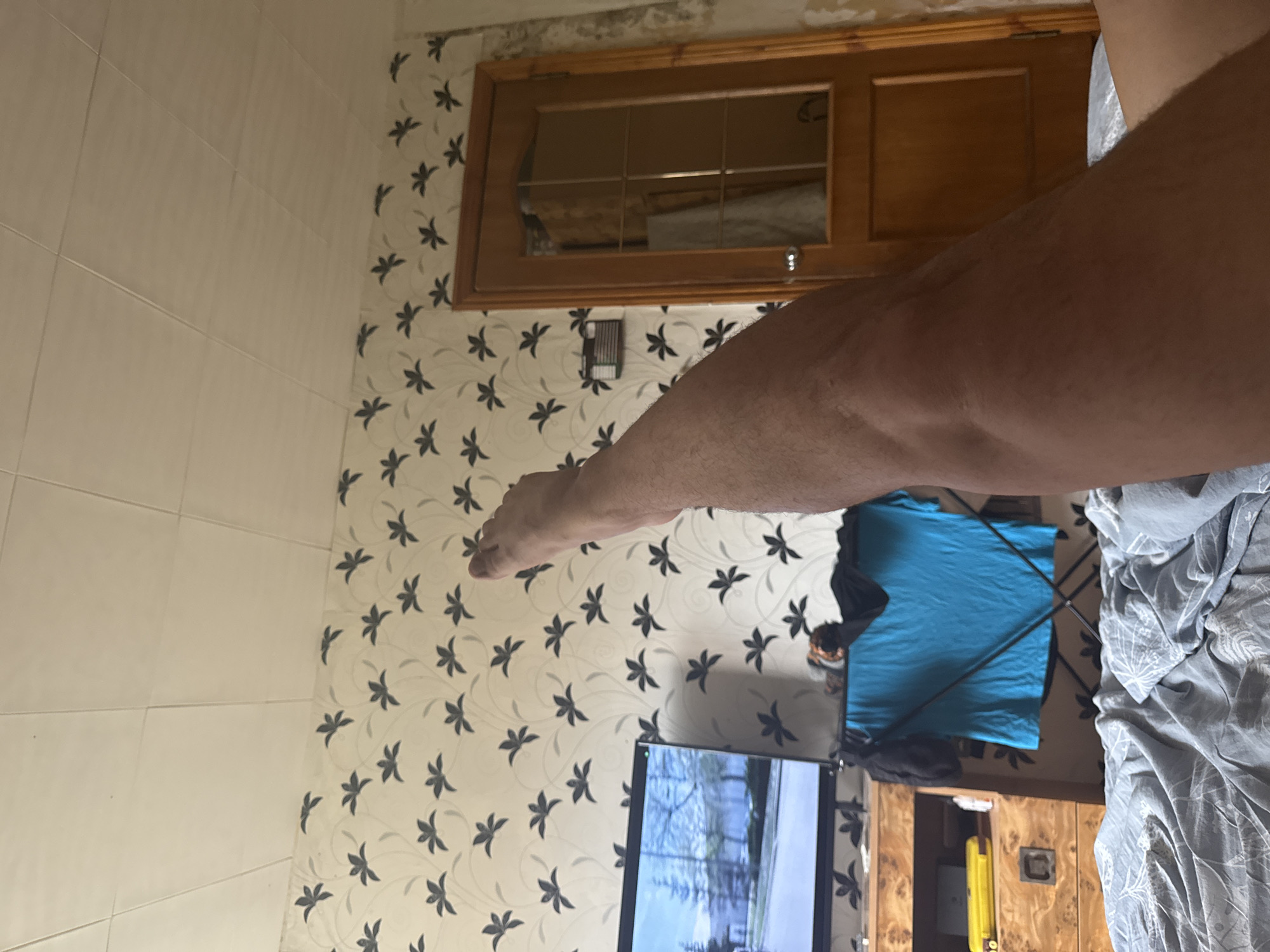 The image captures a bedroom interior in Shcherbinka, Russia. A person's right leg is prominently featured, extended upwards and occupying the foreground. The leg appears to be unshaven and is positioned against a backdrop of floral patterned wallpaper. To the left of the leg, a television screen displays an outdoor scene, possibly a video game or a movie. Below the television, a wooden dresser with a yellow object on its surface and a dark drawer is visible. A drying rack with a blue t-shirt and a black garment is positioned behind the dresser. The room is furnished with a bed covered in grey bedding, patterned with a white design. To the right, a wooden door with a glass panel stands against the wallpapered wall. The ceiling is tiled with white square panels. The overall impression is of a lived-in domestic space.