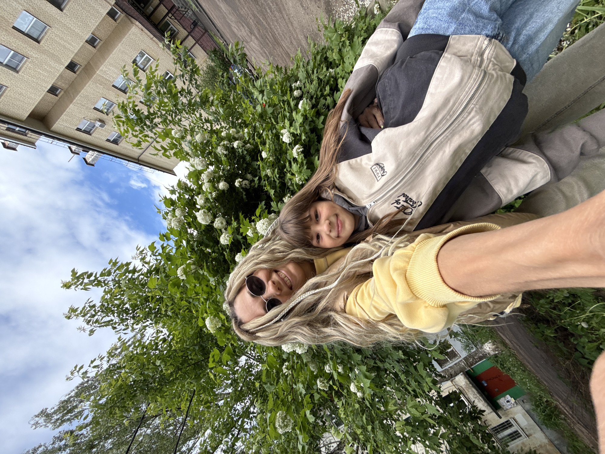 The image shows a selfie taken by a woman with long blonde hair and braids, wearing sunglasses and a yellow sweatshirt. She is lying down with a young girl nestled against her. The girl has long brown hair and is wearing a grey jacket over a grey hoodie. The background consists of a large bush with white flowers, a building with multiple windows under a blue, cloudy sky, and glimpses of other buildings. The location is in the city of Unknown, Unknown. The selfie perspective suggests an interaction of closeness and affection between the woman and the child.