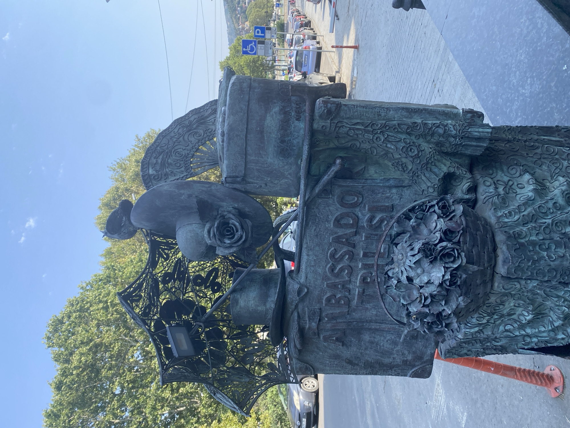 This is a close-up, eye-level shot of a bronze sculpture in Tbilisi, Georgia, depicting a collection of objects. The sculpture features a basket of metal roses at its base, sitting on a textured pedestal. Above the basket, a large, rectangular plaque displays the words "AMBASSADOR" and "TBILISI" in raised lettering. Resting on top of this plaque are a man's top hat and a decorative umbrella made of intricate metalwork, which has a modern spotlight attached to it. Behind the umbrella, a detailed bronze fan and what appears to be a hatbox are visible. A large, sculpted rose is placed on top of the hatbox. To the right of the sculpture, a street scene unfolds with parked cars, a blue disabled parking sign, and a P parking sign. In the background, lush green trees and a hilly cityscape are discernible under a clear blue sky. The sculpture is located on a paved sidewalk adjacent to a street.