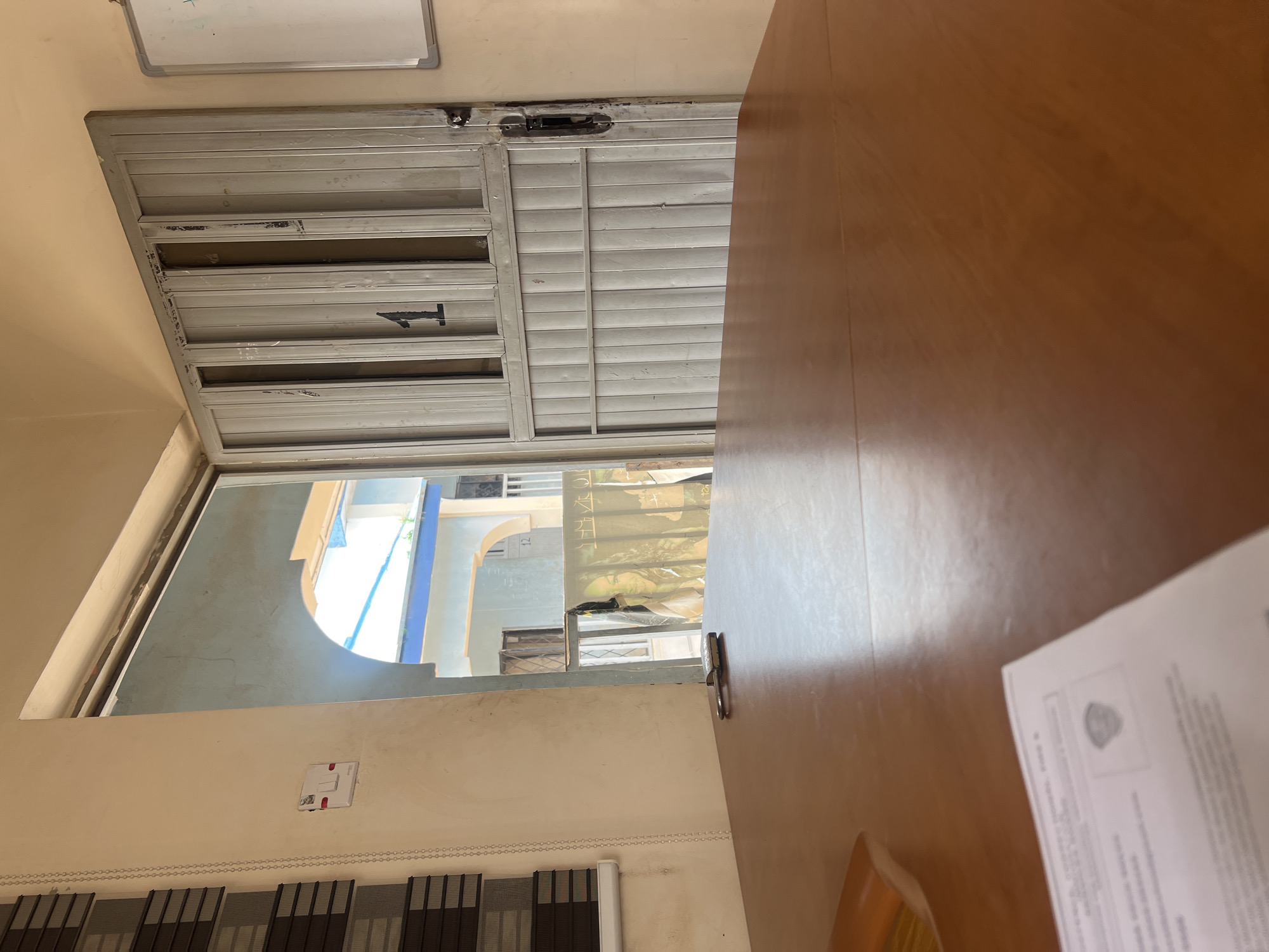 The image depicts an interior view looking towards an open doorway. In the foreground, a wooden table with a glossy surface dominates the lower half of the frame. A document with text and an emblem is partially visible on the table in the bottom left corner. A silver lock is placed on the table towards the right side of the foreground.
The doorway leads to an outdoor area. Through the doorway, a metal gate or door is visible, painted a light gray color and featuring vertical slats. The number "1" is prominently painted in black on this gate. To the left of the doorway, a wall is visible with a white electrical outlet and a chain hanging down. Further outside, through the doorway and the gate, a building with blue and yellow accents and an arched entrance is observed, suggesting a residential or commercial setting. A poster featuring images of women is partially visible hanging outside. The environment appears to be in a city, and the overall impression is of a functional, somewhat aged, urban setting. The scene is consistent with observations of typical street-level architecture and security measures found in cities like Jos, Nigeria, characterized by multi-story buildings, security gates, and exposed electrical fixtures.