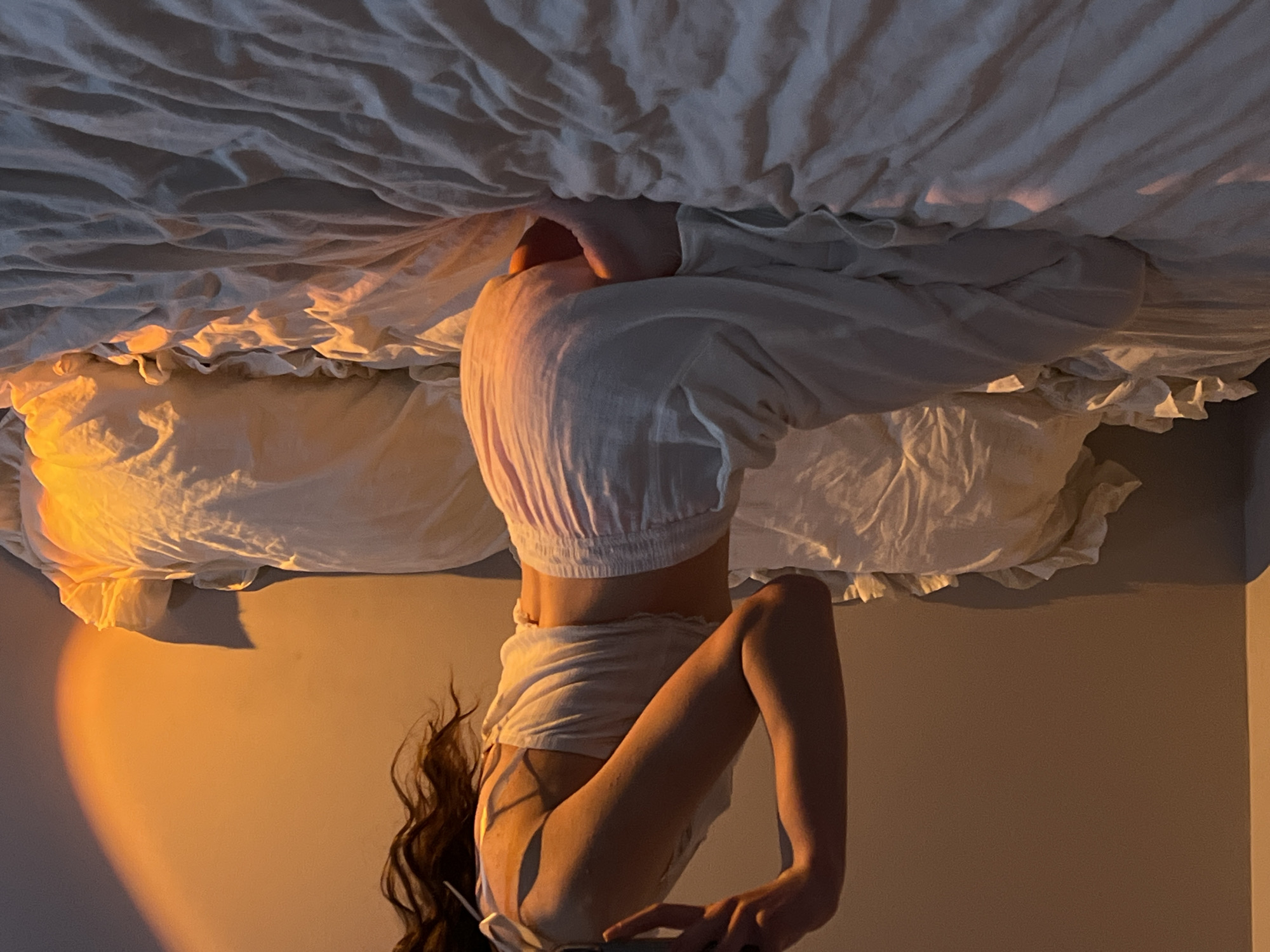 A female is upside down in a room with a bed. She is wearing white clothing. The image is lit with warm-toned lighting. It is unclear if the location is in the city of Ternopil, Ukraine.
