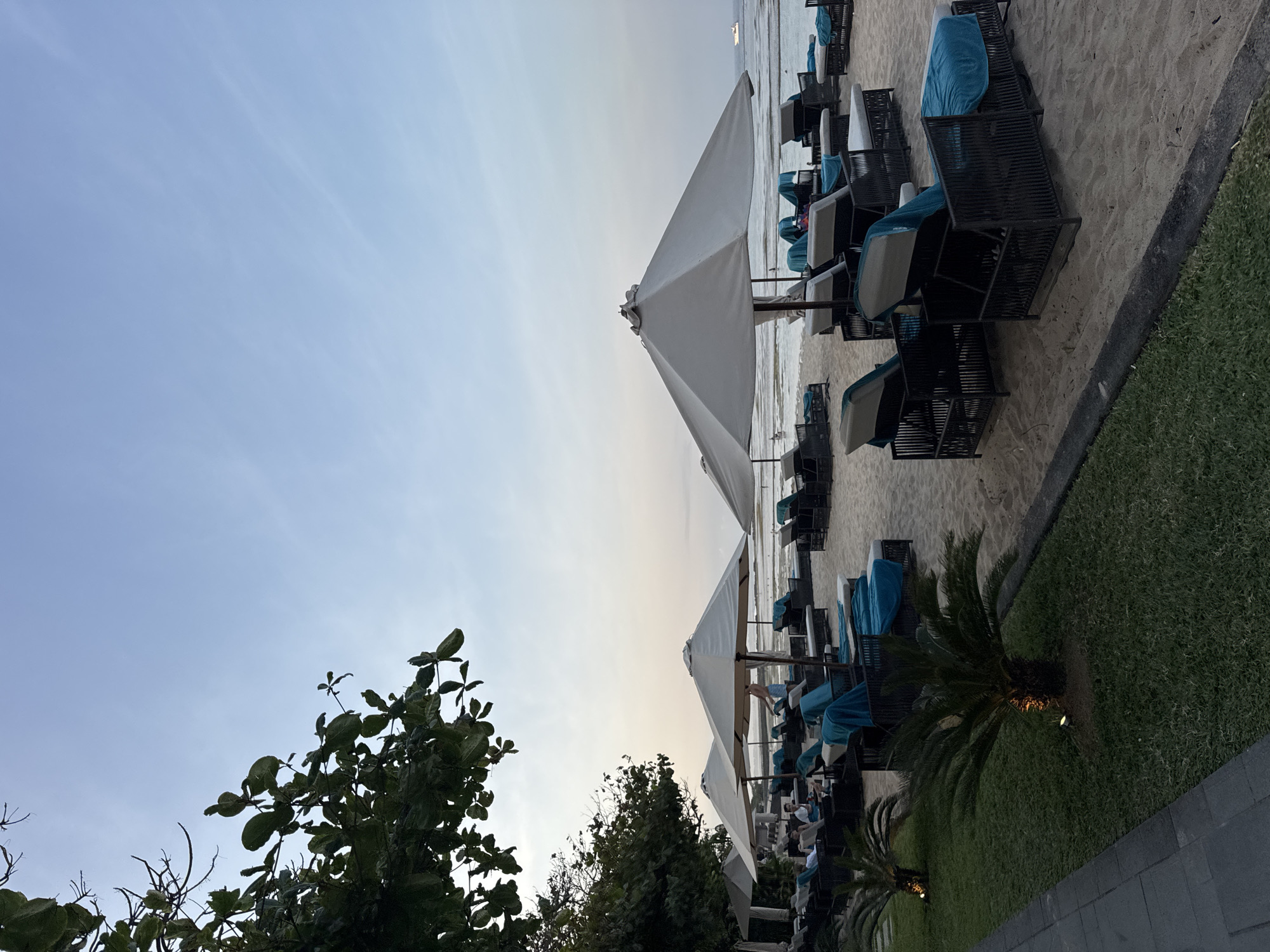 The image shows a beach in Kuta Selatan, Indonesia, with rows of empty lounge chairs covered in blue towels, suggesting it's either early morning or late evening. Large white umbrellas provide shade over some of the chairs. In the background, the ocean is visible, with a small boat in the distance. Lush green trees are on the left side of the frame, casting shadows on the sand. The sky is a soft blue, indicating the time of day. A few people are seen sitting on chairs in the distance.