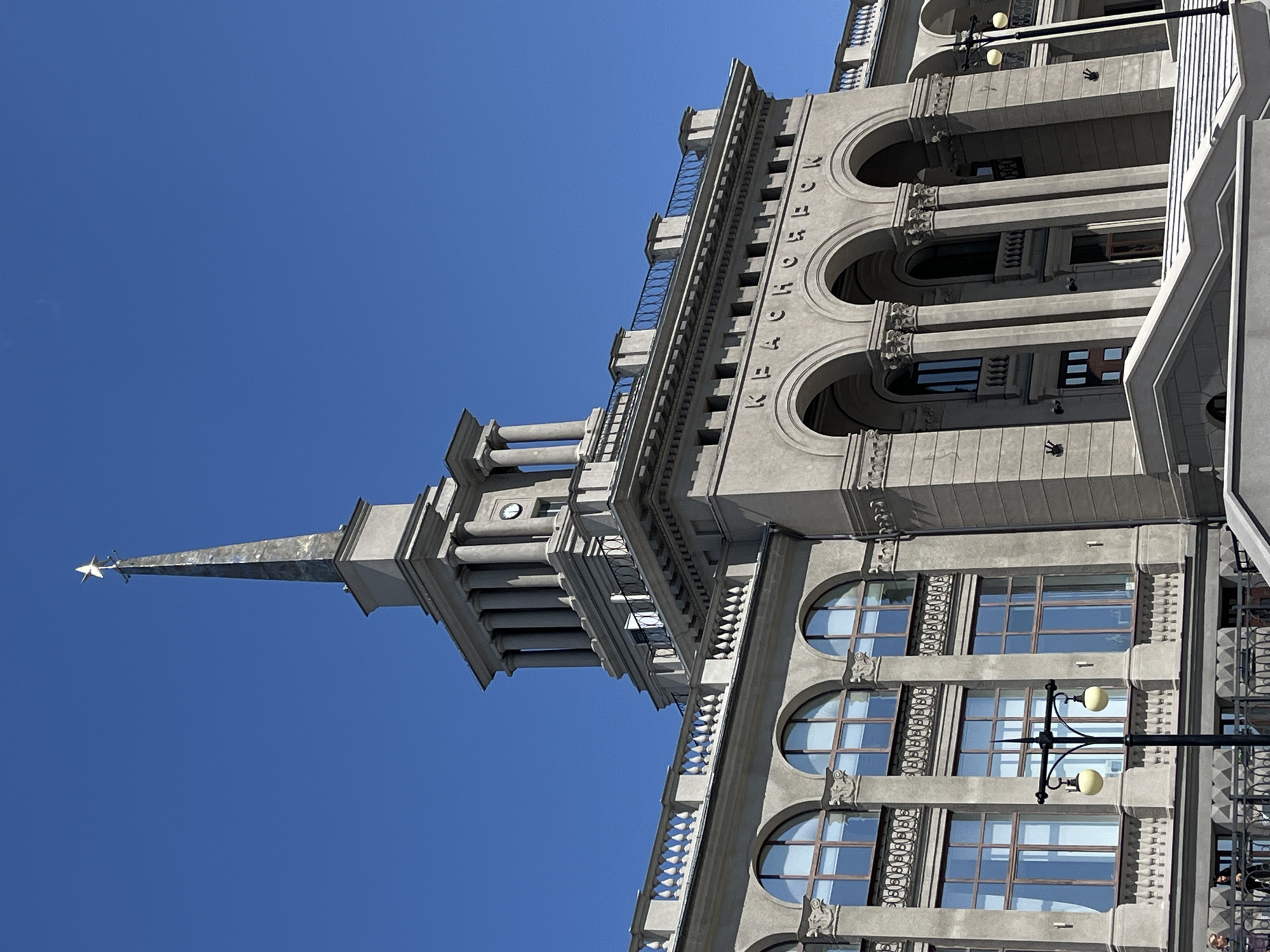 A low-angle shot depicts the architectural details of a building in Krasnoyarsk, Russia, against a clear blue sky. The building features a spire topped with a star, classic column designs, arched windows, and decorative molding. The name "Krasnoyarsk" is vertically inscribed along the building's facade in Russian. Lamp posts are visible in front of the building, and a few people are barely perceptible on what appears to be a balcony or sidewalk.
