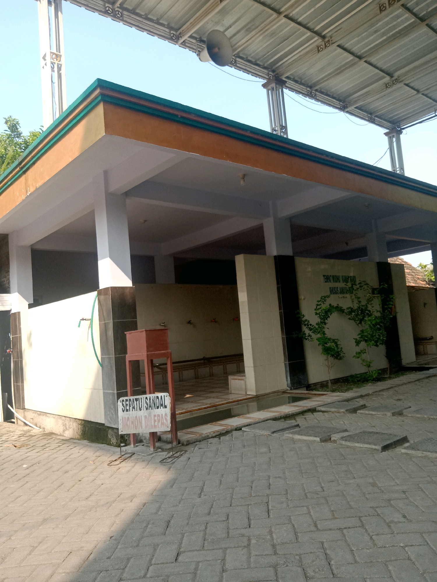 This image depicts an outdoor area in Sugihwaras, Indonesia, featuring a building with an open-air covered structure. The structure has a prominent roof supported by columns and appears to be a place for ablution or washing, as indicated by the presence of faucets and a sign that reads "SEPATU/SANDAL MOHON DILEPAS" (Shoes/Sandals Please Remove). The ground is paved with interlocking bricks. In the background, there is a wall with text, a small tree, and more of the covered structure. The sky is clear and blue. The scene appears to be clean and well-maintained. There are no people visible in the image.