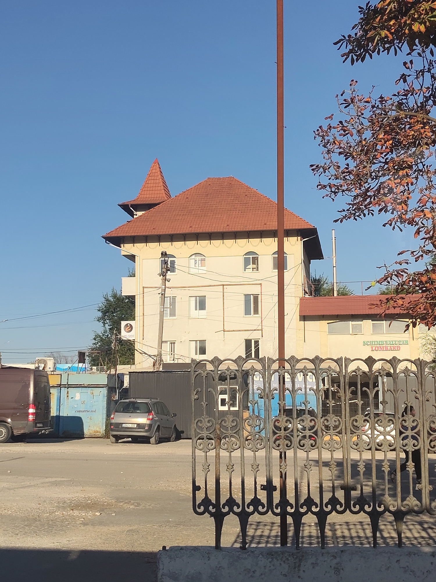A multi-story building with a red tiled roof is visible against a clear blue sky. A pointed turret tops one section of the building. The building is cream-colored with several windows of various shapes and sizes. A sign on the building reads "SCHIMBVALUTAR LOMBARD" in green lettering. In the foreground, an ornate metal fence with decorative scrollwork obscures a street scene. Behind the fence, a silver car is parked on a paved surface. To the left of the car, a dark-colored van is partially visible. Several industrial-style containers are also present in the background. The overall setting appears to be an urban environment. The image was taken in Chisinau, Moldova.