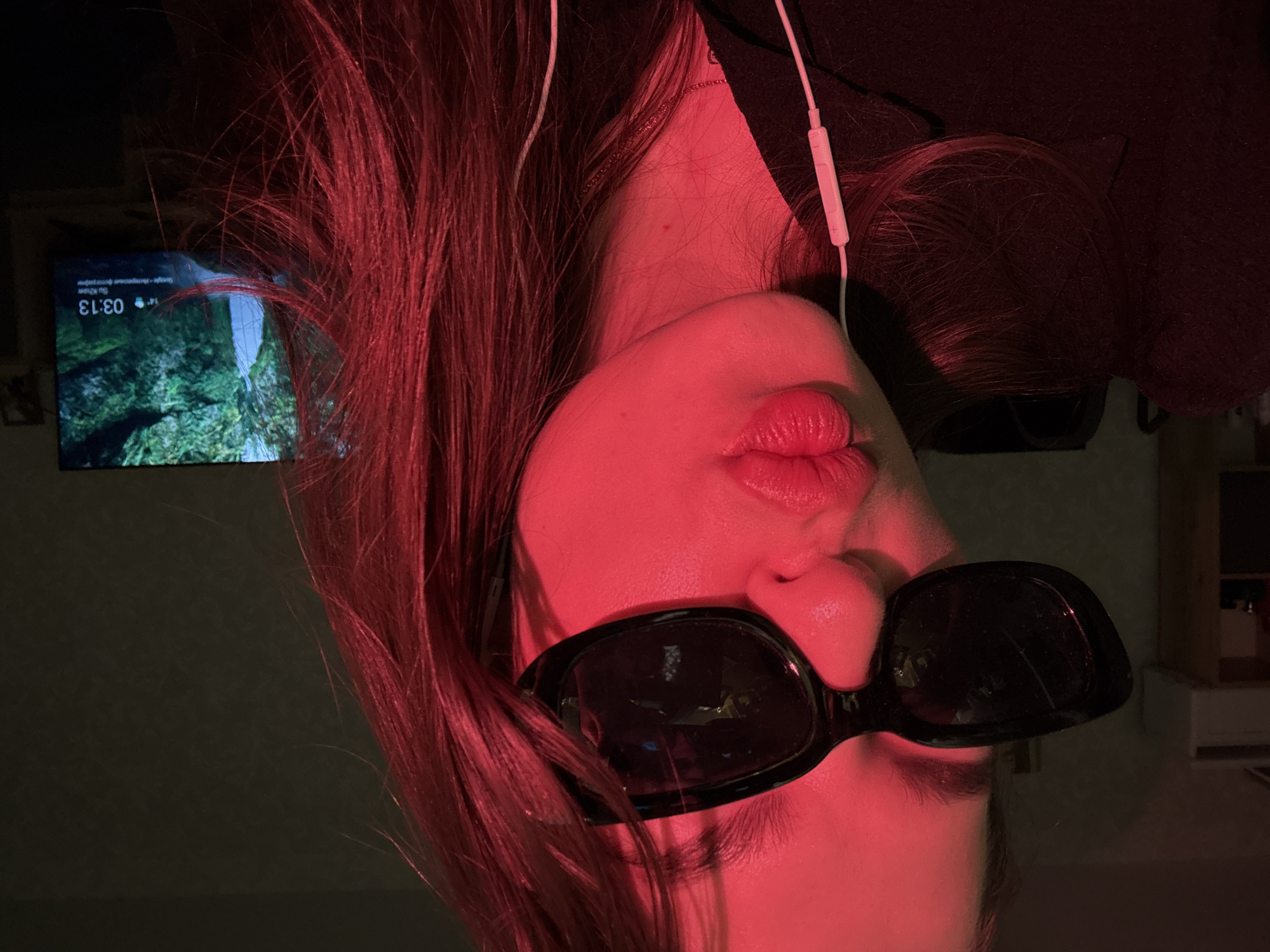 A photograph taken indoors in what appears to be Chaikovsky, Russia shows a woman with red hair and black sunglasses upside down. She is wearing headphones with a white cord. In the background, a television screen displays a scenic view, with a time reading "03:13". The overall lighting is red.
