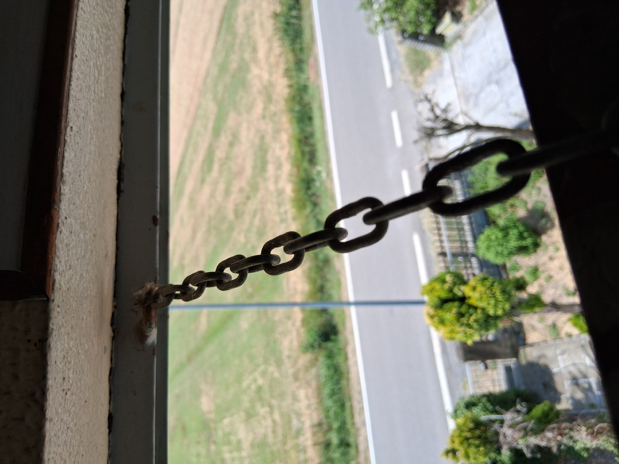 An exterior shot looking down at a hanging metal chain suspended from a building's overhang. The chain is the primary subject, with its links clearly visible. In the background, a rural landscape is visible, featuring a road, green fields, and some trees. The scene appears to be located in Colorno, Italy, possibly on a balcony or elevated structure overlooking the outdoors.