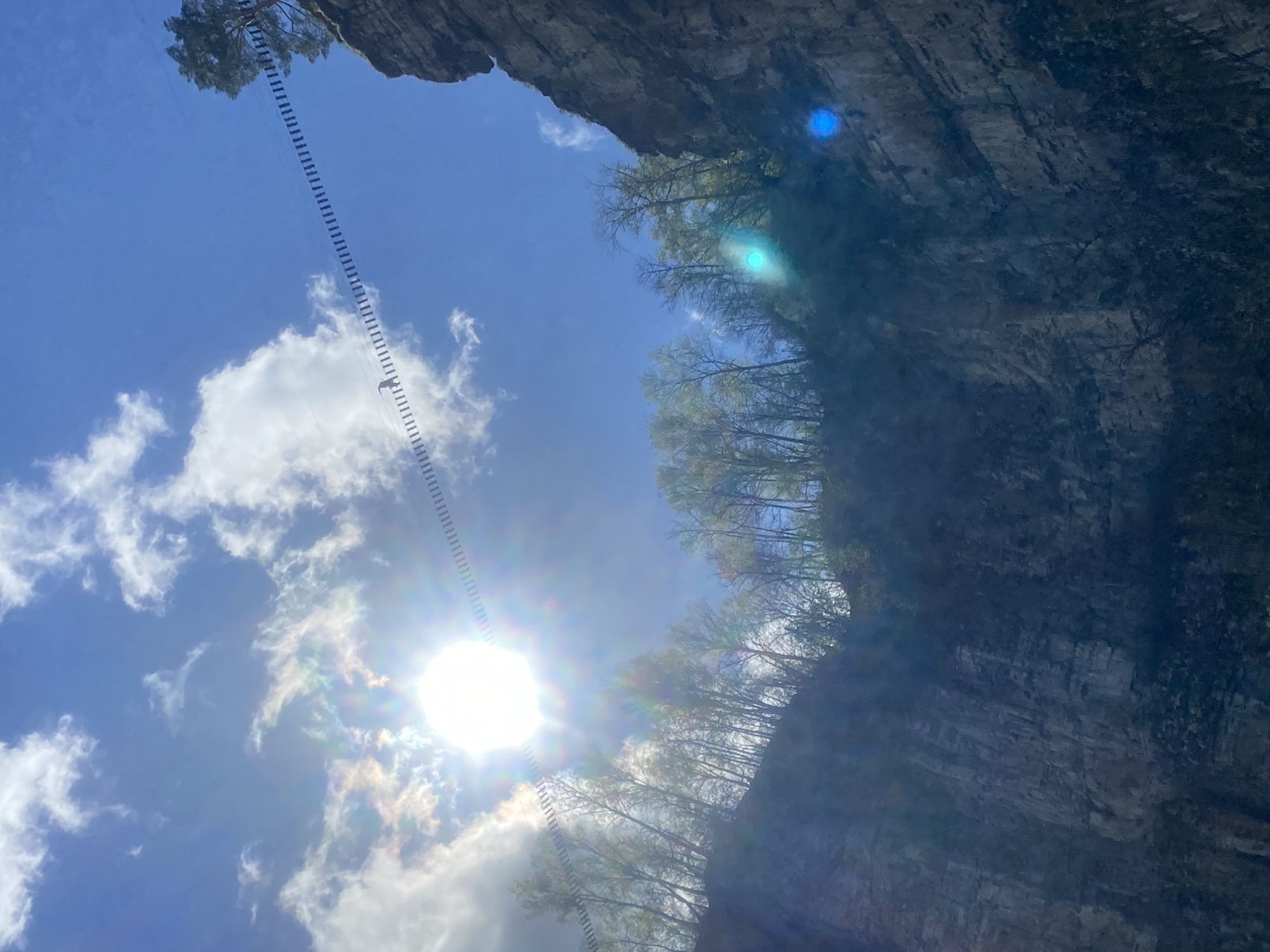 The image depicts a view from below, looking up at a rocky cliff face and a bright, sunlit sky. A suspension bridge or walkway with a linear pattern of platforms extends from the top of the cliff into the sky. Trees are visible along the top edge of the cliff. The environment consists of a rock formation, trees, and the sky with clouds. The location is Kul'metovo, Russia, based on the user's request.