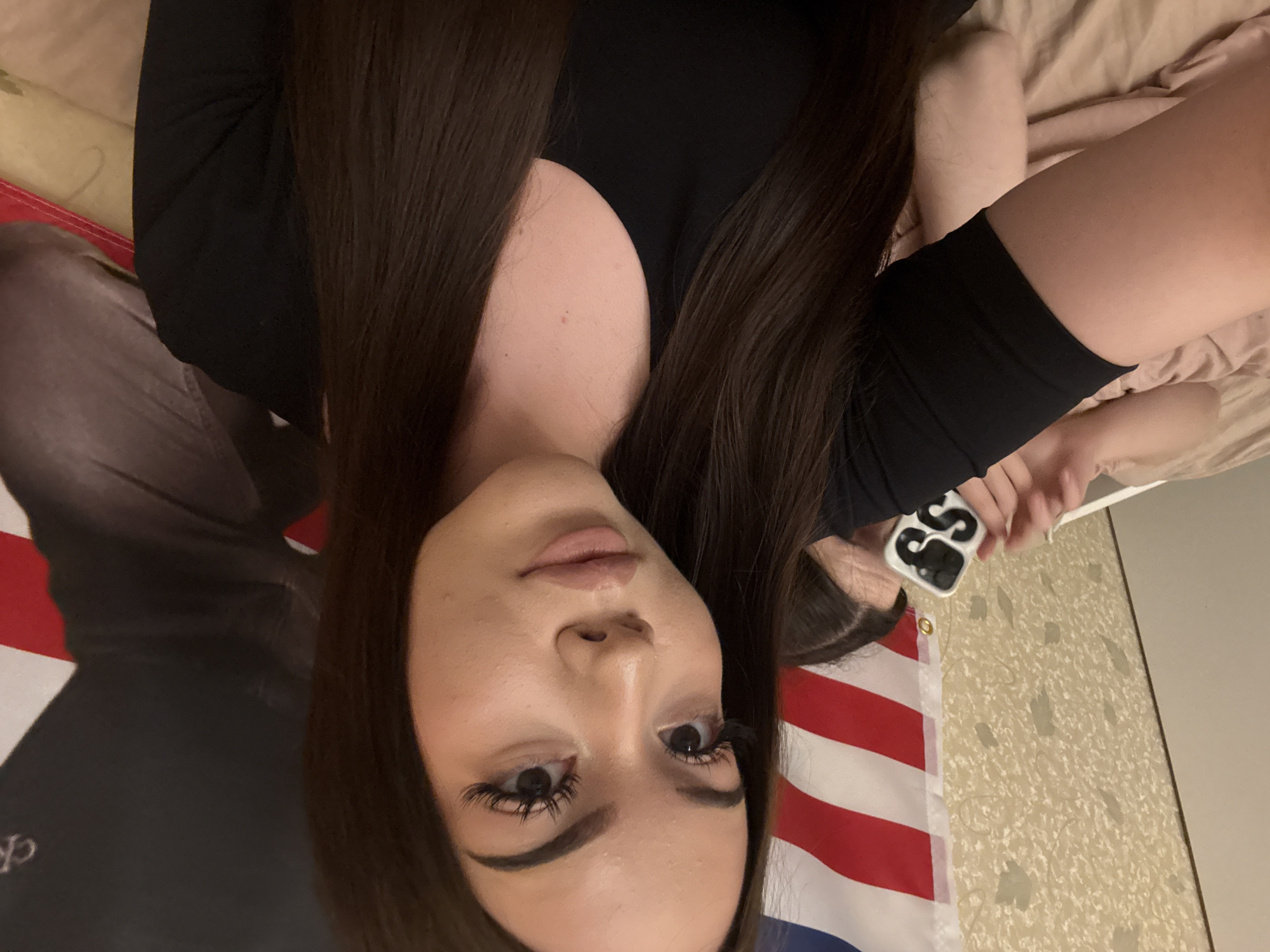 An image, rotated 90 degrees clockwise, captures a young woman lying down indoors, gazing directly at the camera. She has long, dark brown hair, dark, well-defined eyebrows, and pronounced eyelashes. Her lips are full and light-colored, and her facial expression is neutral.
She is wearing a black, long-sleeved top or dress. Her right arm is visible, holding a smartphone with a white case featuring a distinctive black "S" logo or design. A portion of an American flag, characterized by red and white stripes and a blue section with white stars, is visible beneath her on the left side of the frame. The background includes light brown or tan bedding in the upper and lower right sections, and a light-colored wall with a subtle, textured, possibly leaf-like pattern on the far right. The lighting suggests a well-lit indoor environment. The image is set in Naberezhnye Chelny, Russia.