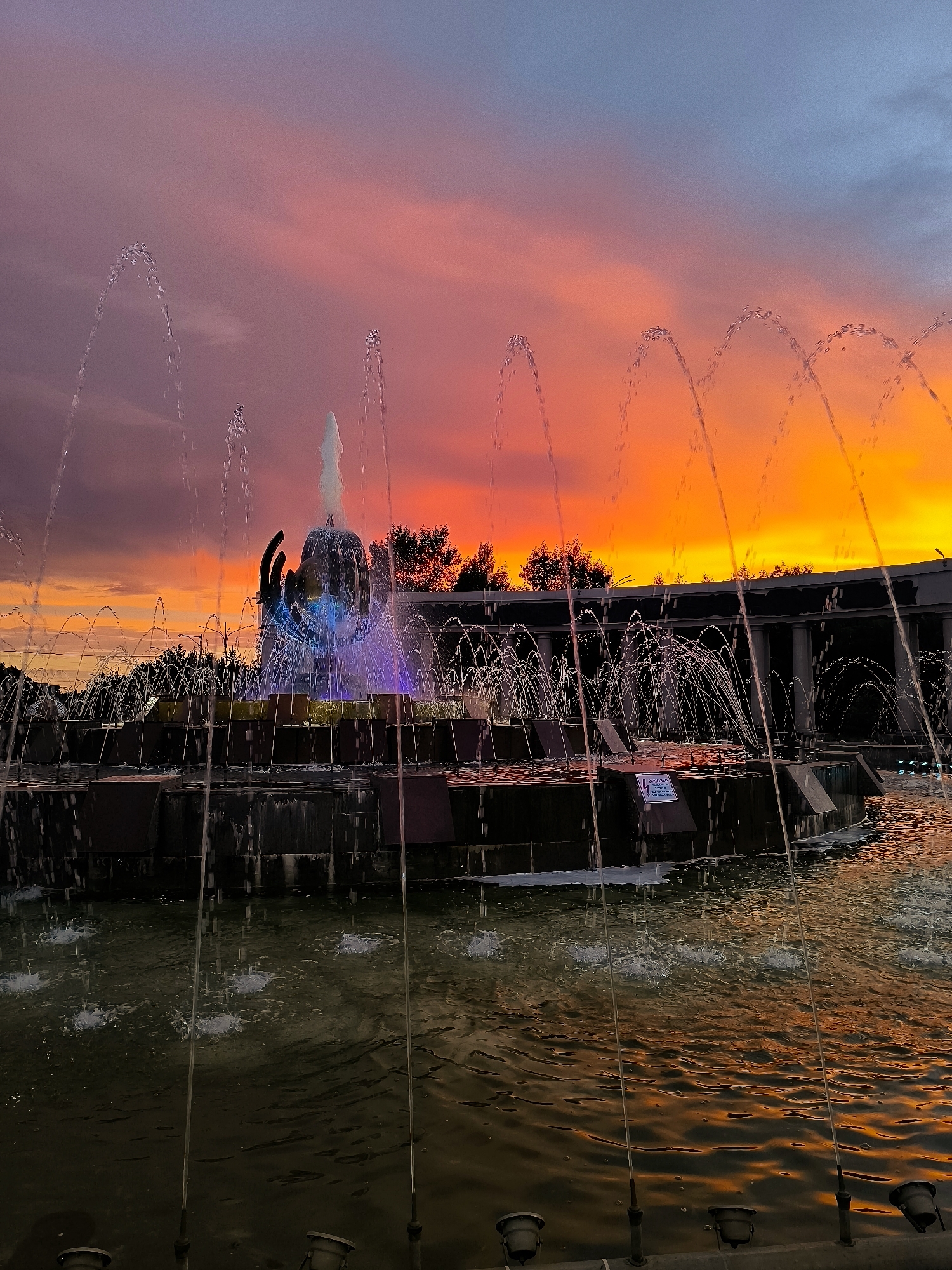 The media file depicts a large, multi-tiered fountain at dusk. The fountain features numerous water jets, some forming graceful arcs, others creating spray. The central structure of the fountain is illuminated with vibrant blue and purple lights, highlighting its sculptural elements. The water in the fountain reflects the dramatic colors of the sunset, creating an orange and pink glow on its surface.
The sky is a gradient of colors, transitioning from a deep purple and pink in the upper portion to a brilliant orange and yellow near the horizon, indicative of a sunset. Silhouetted trees and a long, arched building with columns can be seen behind the fountain, suggesting an urban park or public square setting. The overall atmosphere is serene and visually striking, amplified by the intense colors of the sky and the illuminated fountain.
There are no people visible in the media file. The time of day is clearly evening, during sunset. The weather appears clear, with the vibrant sky suggesting fair conditions. A small, white sign with black text is visible on one of the fountain's tiers, but the text is too small to be legible. The location is identified as Qarağandı city administration, Kazakhstan.