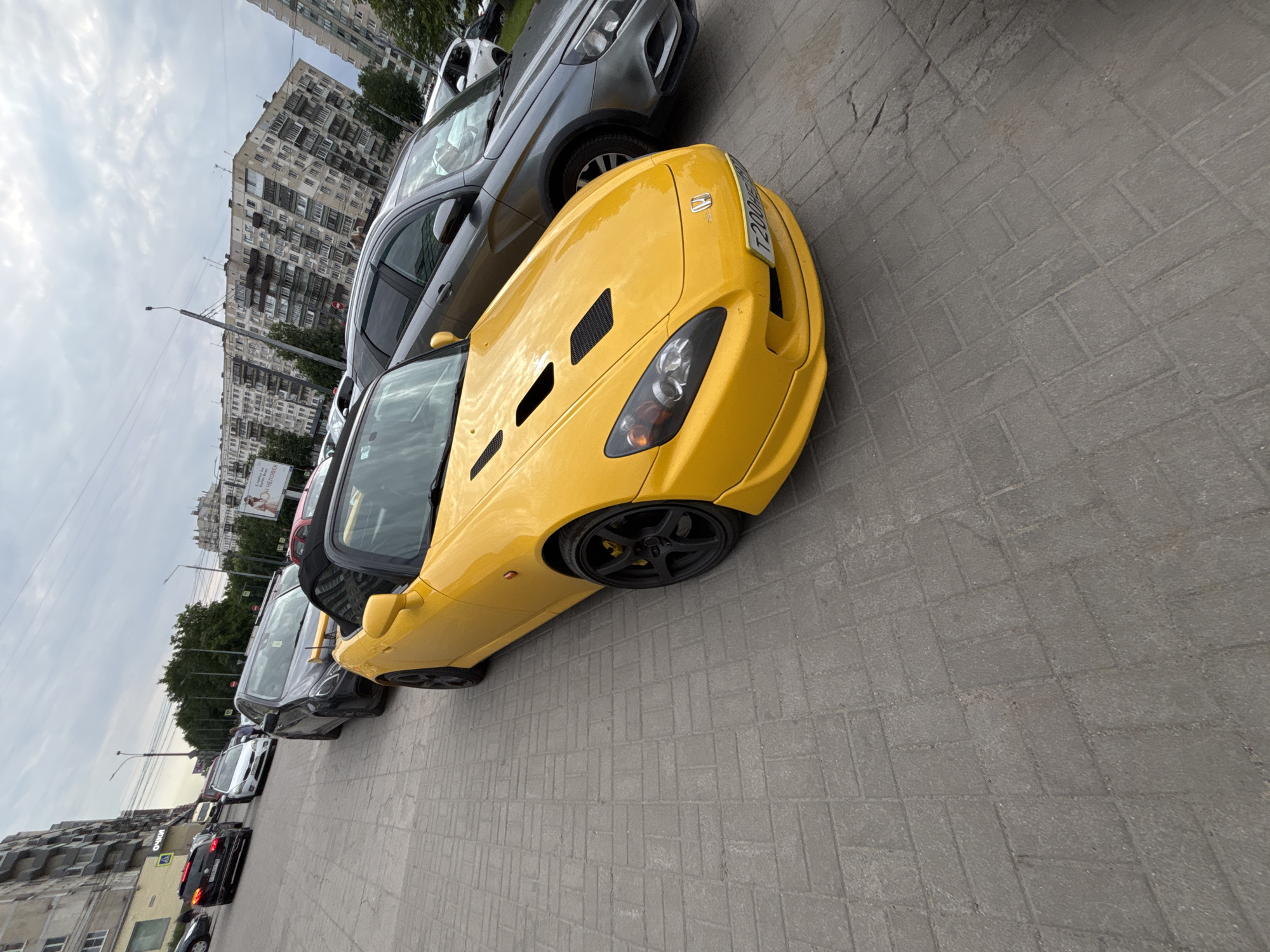 A yellow Honda S2000 convertible is parked on a sidewalk made of gray pavers, next to other cars and apartment buildings in Ladozhskoe Ozero, Russia. The S2000 faces towards the right, its front bumper and hood visible. To its right, a gray sedan is parked. In the background, several other cars are lined up, and large, multi-story apartment buildings line the street. The sky is overcast.